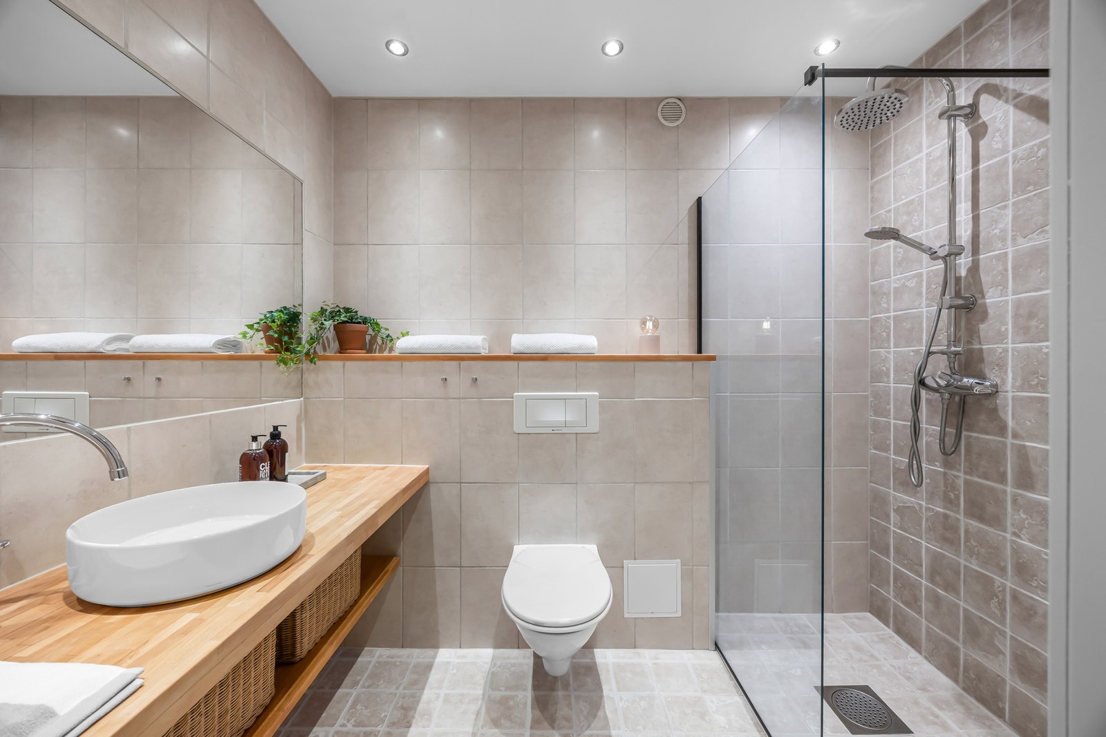 Bad med dusj, vegghengt toalett, servant på benk og stor speilflate. Fliser på vegger og gulv. Galleribilde