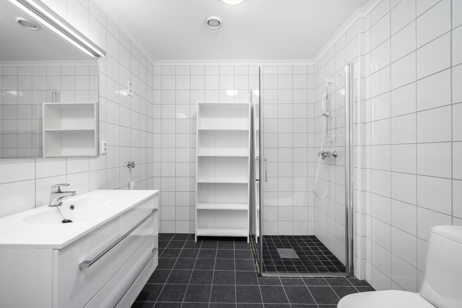 Pent flislagt bad på 5 kvm med toalett, dusjnisje med dører i herdet glass og innredning med heldekkende hvit vask, underskuffer med slette fronter og speil med lys. Varmekabler, opplegg for vaskemaskin og avtrekk via balansert boligventilasjon. Galleribilde