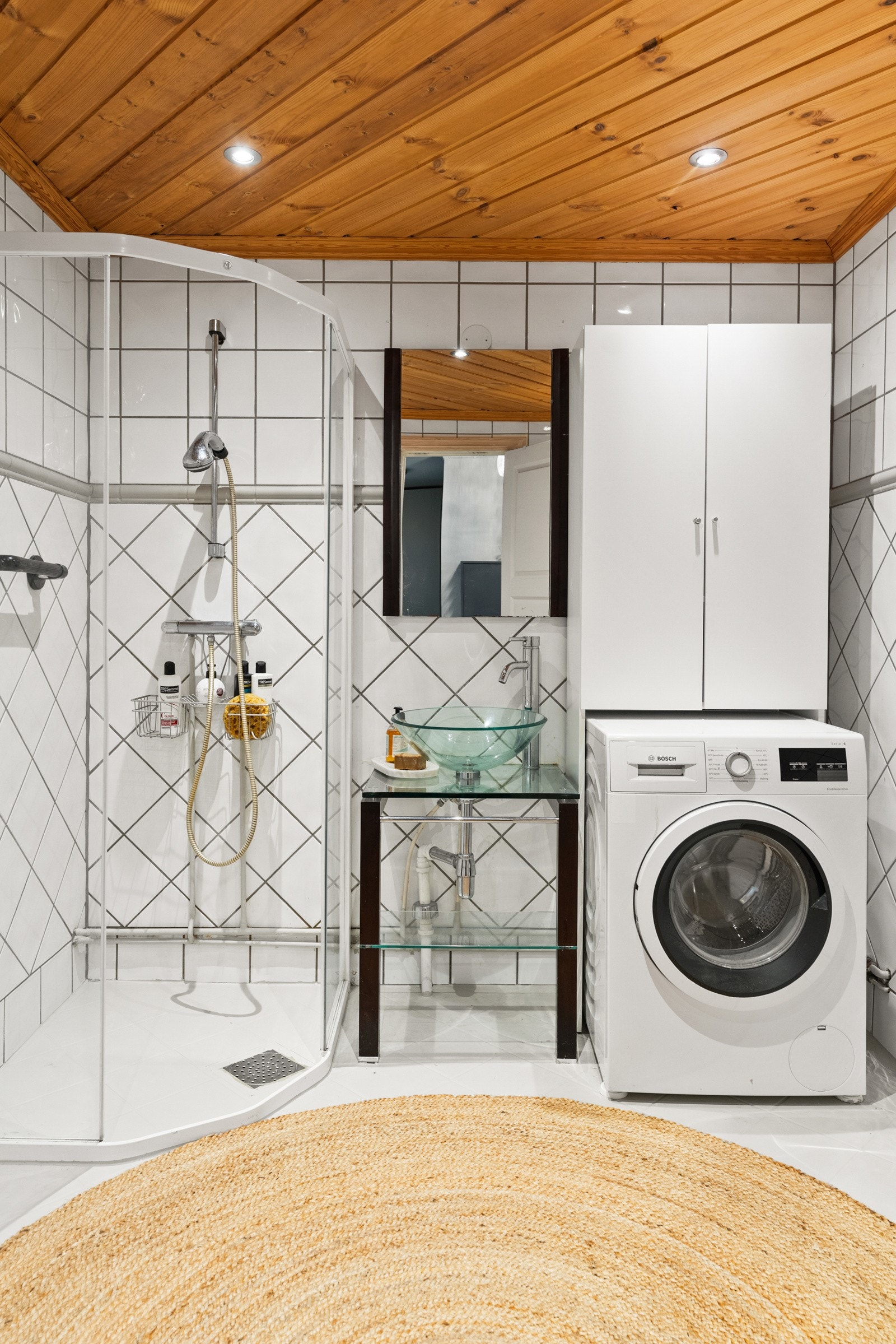 Rommet er innredet med baderomsmøbel med hylle samt en servant i glass, vegghengt skap, dusjvegger/ hjørne, og opplegg for vaskemaskin Galleribilde