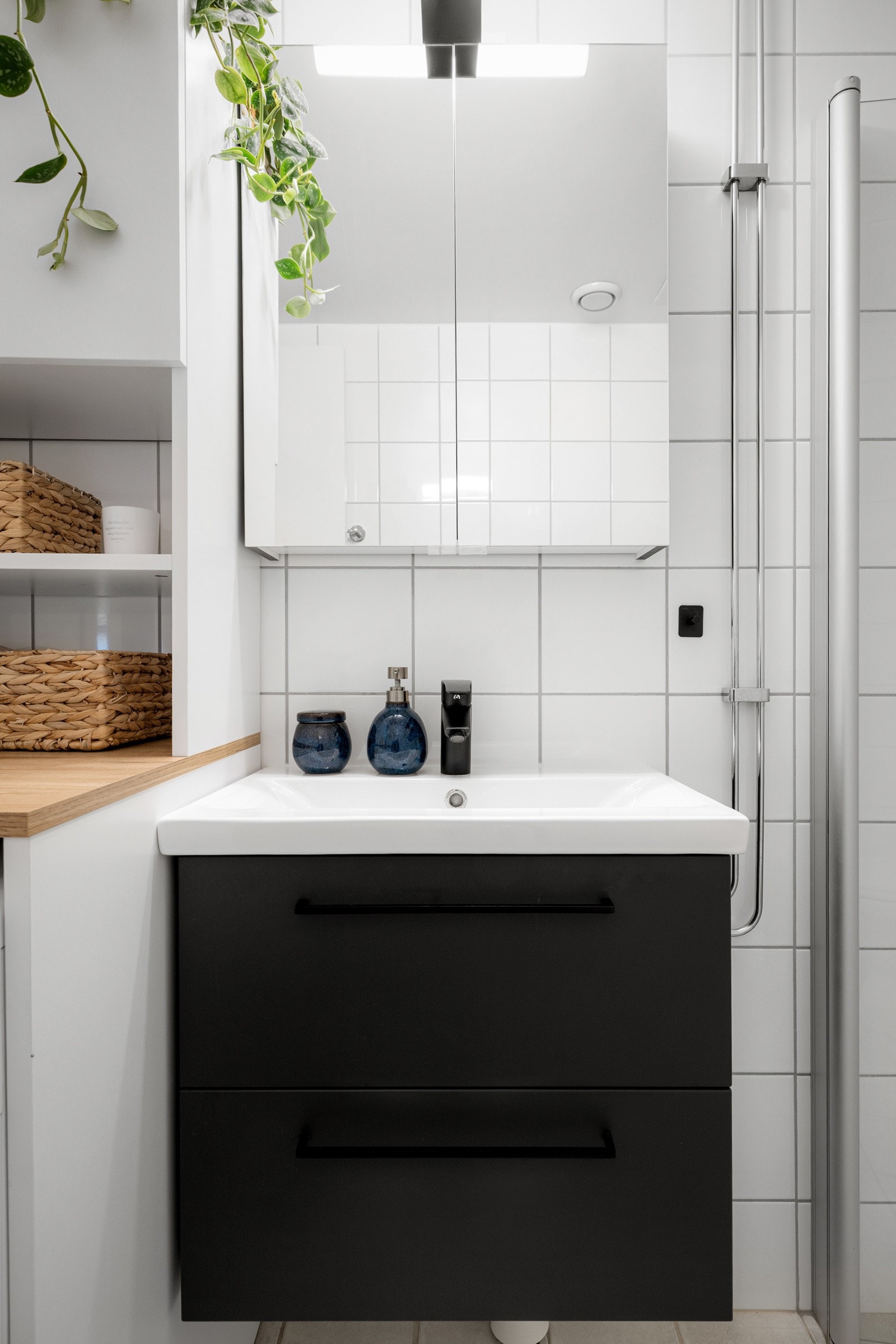 Moderne baderomsinnredning med skuffer og heldekkende servant med sort armatur, speilskap med belysning over. Baderommet er også utstyrt med dusjhjørne med innfellbare glassdører og gulvstående toalett. Galleribilde