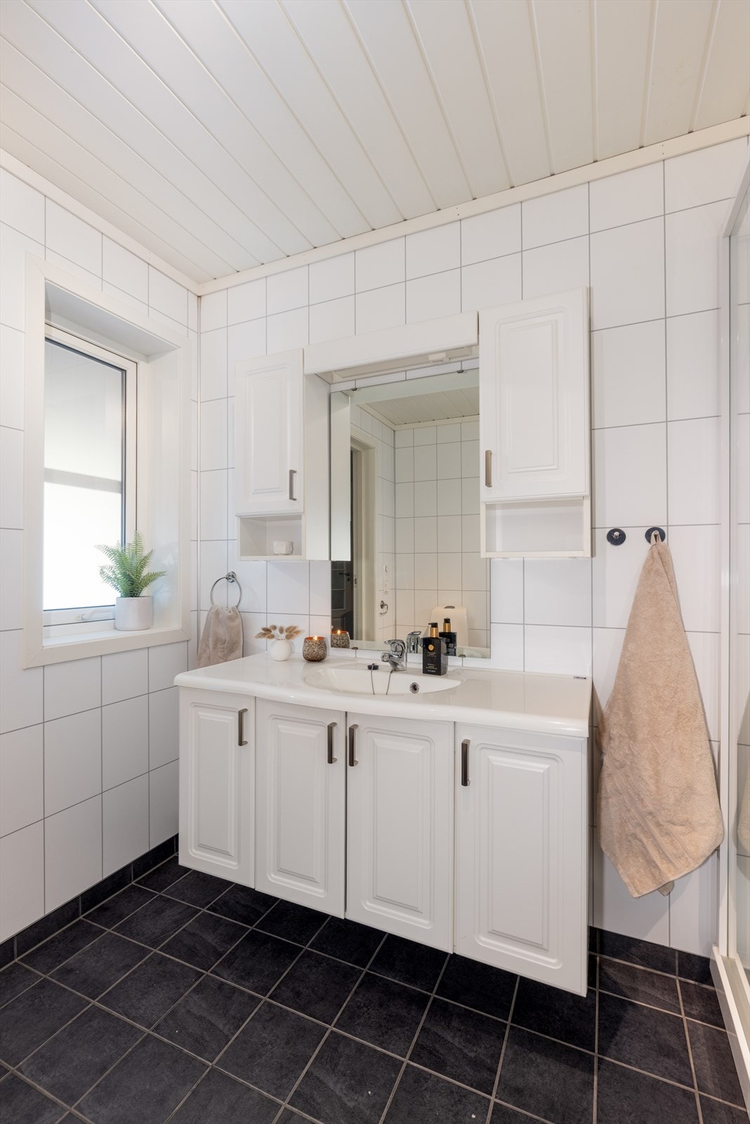 Romslig baderomsmøblement med under- og overskap, heldekkende servantplate og speil og belysning over vask. Videre er det dusjkabinett i hjørnet, et veggmontert høyskap og opplegg og plass for vaskemaskin og tørketrommel. Galleribilde