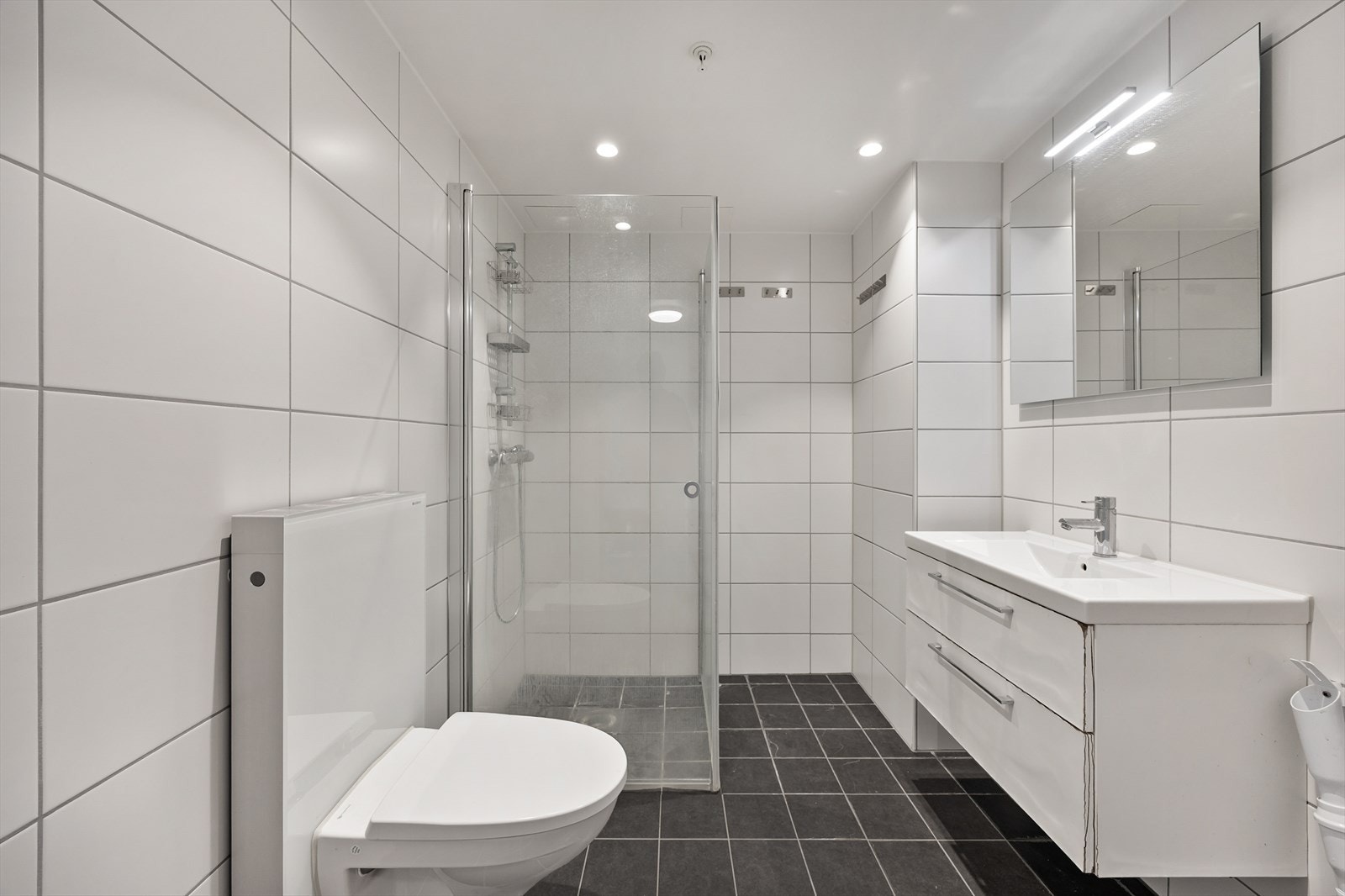 Badet har dusjnisje med glassdører, vegghengt toalett og varmekabler i gulvet. Galleribilde