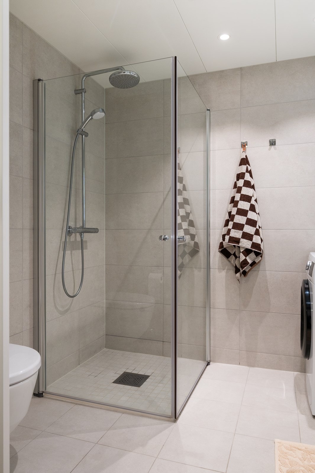 Dusjhjørnet er rett på gulvet med nedfliset dusjsone, innfellbare rette glassdører, dusjbatteri, regndusj og hånddusj - en moderne og praktisk løsning Galleribilde