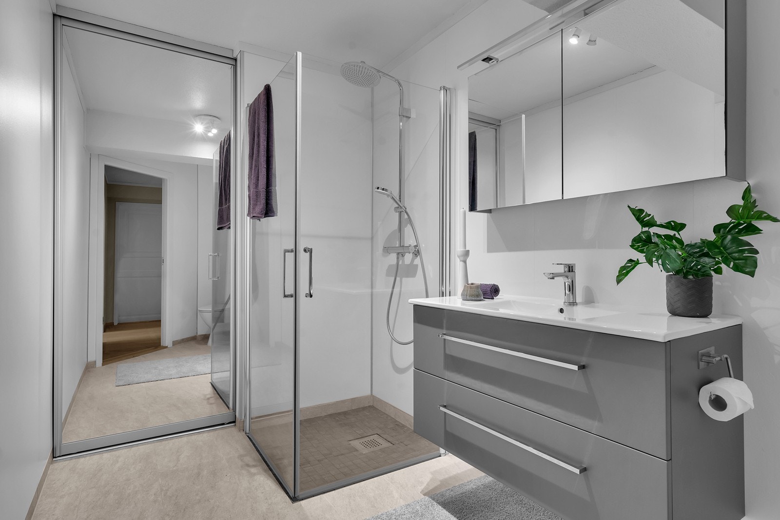 Baderomsinnredning med vask og overskap med speil, dusjnisje med glassvegg og toalett. Vaskeromsavdelingen er skjult bak praktiske skyvedører. Galleribilde