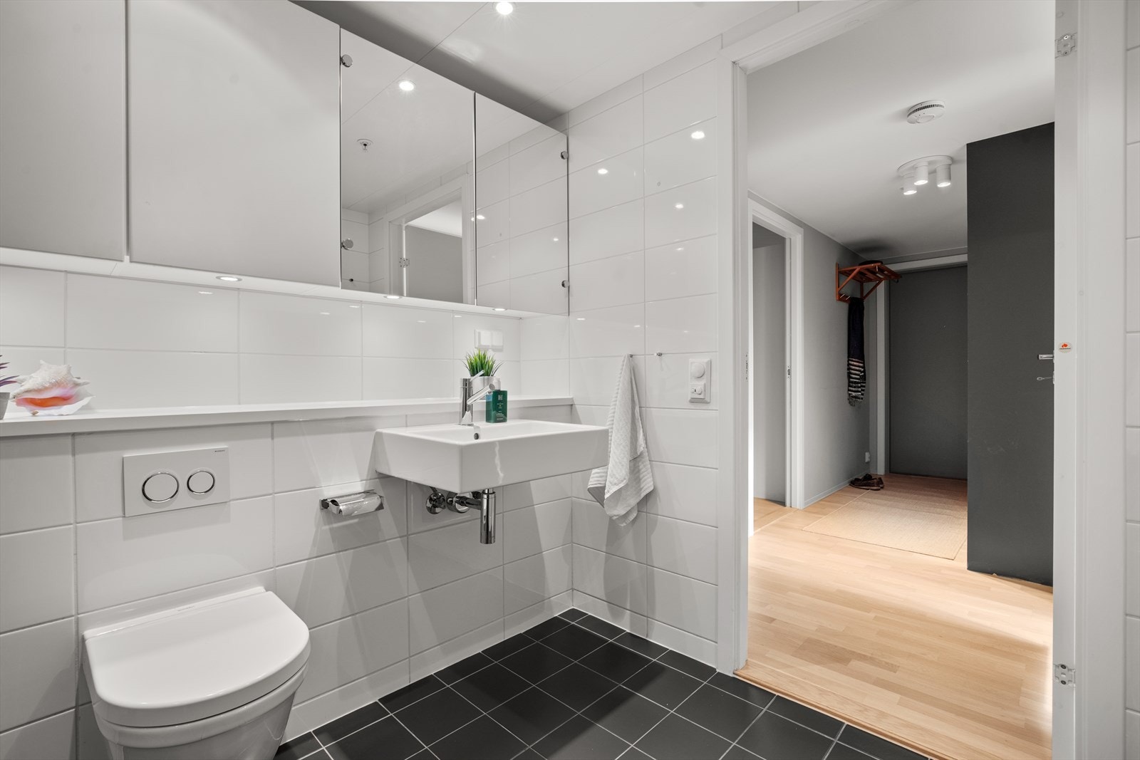 Badet har en praktisk og moderne løsning med veggmontert toalett, servant med speilskap og svingbare dusjvegger ved et nedsenket dusjhjørne. Galleribilde
