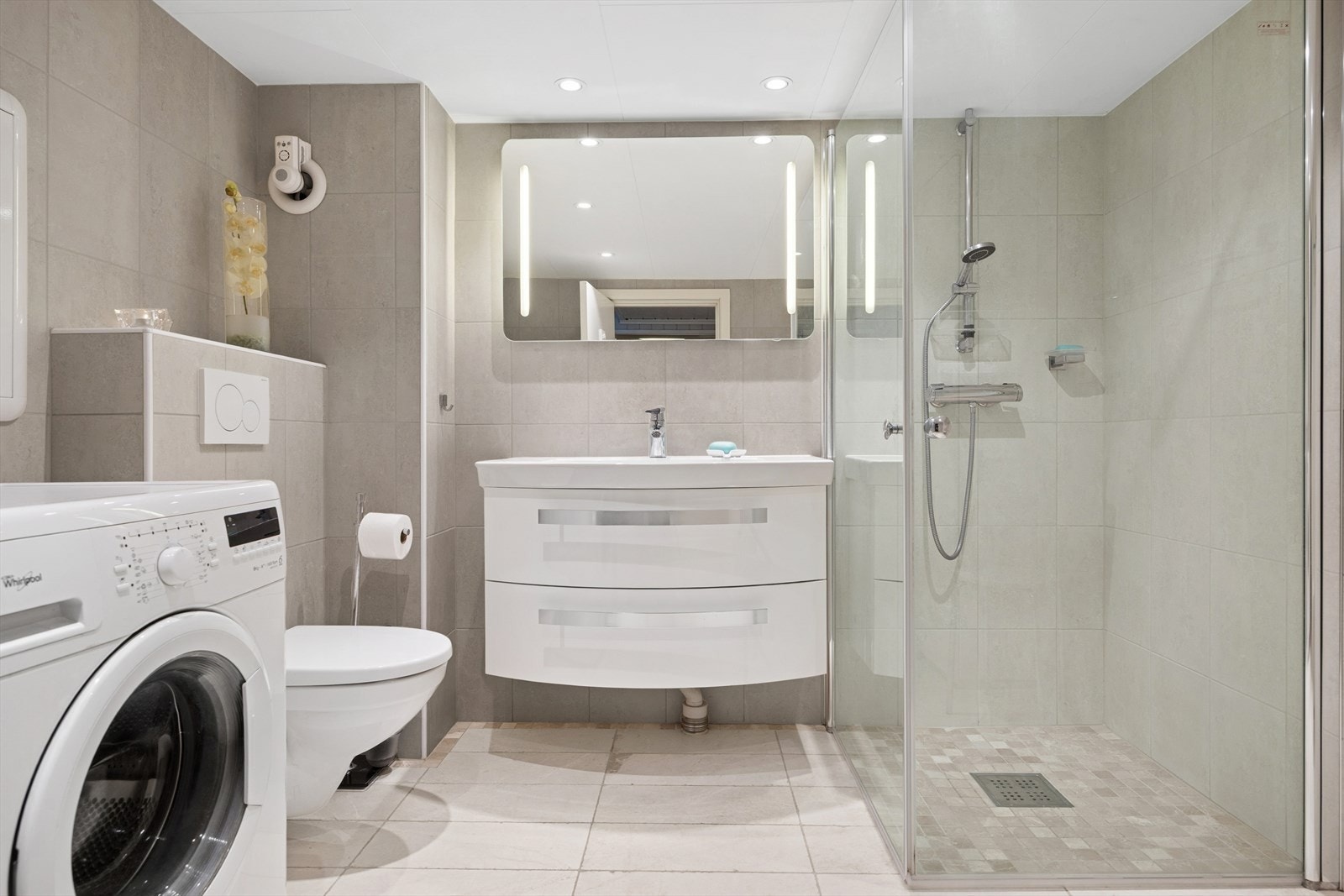 Badet har dusjvegger av glass og dusjgarnityr på vegg, vegghengt toalett, innredning med servant og opplegg for vaskemaskin. Badet opplyses å være oppgradert i 2015. Galleribilde