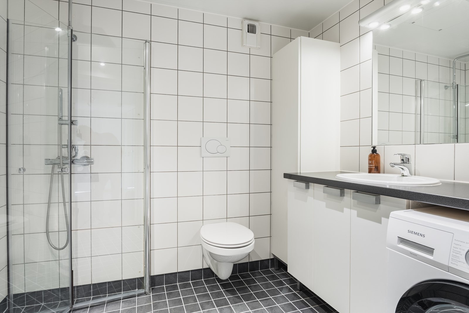 Stort flislagt bad fra byggeår - utstyrt med dusjhjørne, mekanisk avtrekk, opplegg for vaskemaskin og veggmontert wc. Galleribilde