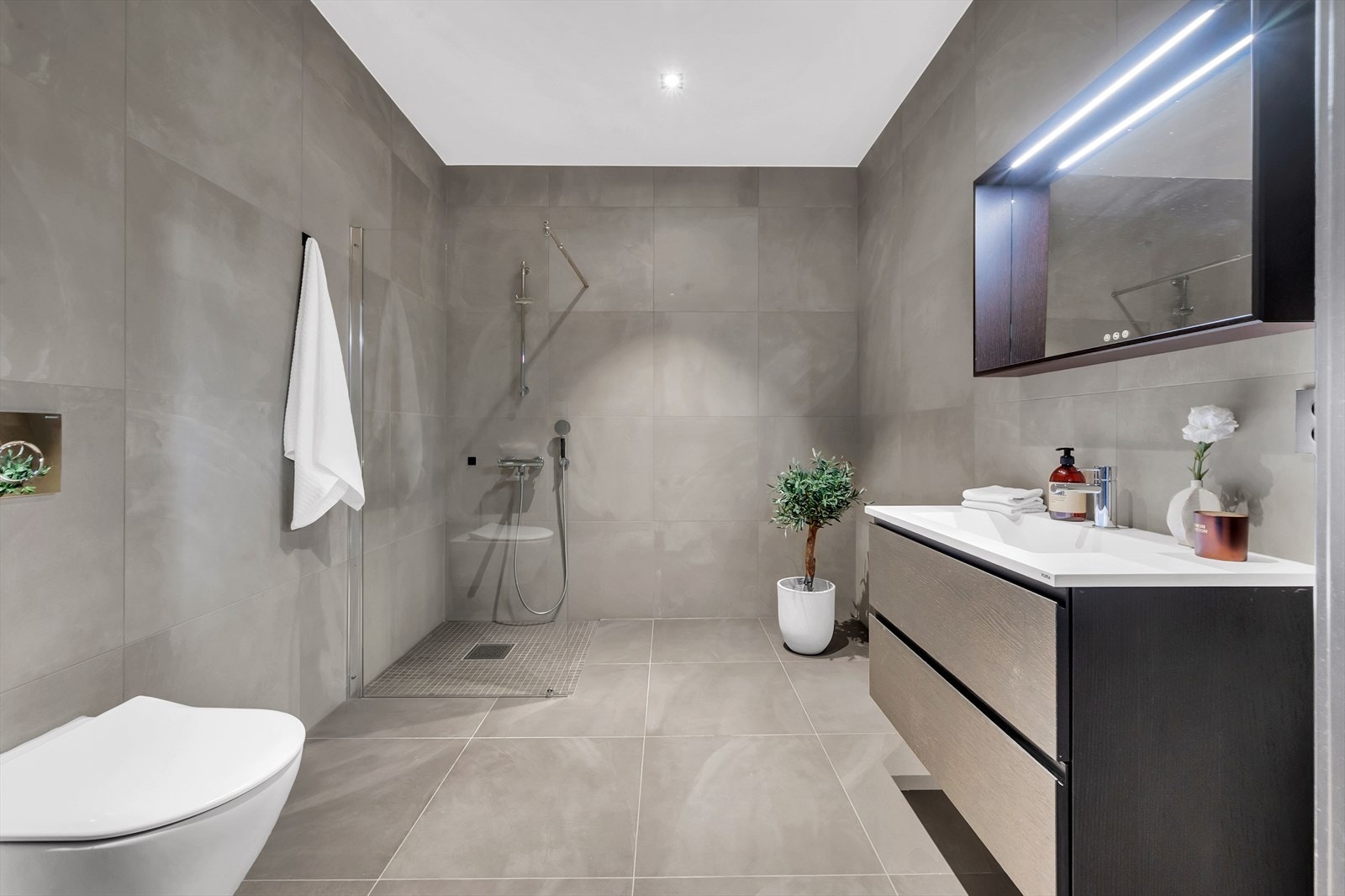 Lekkert flislagt baderom med dusjhjørne, servant med innredning og toalett. Grå fliser på gulv og vegger gir en god romfølelse Galleribilde