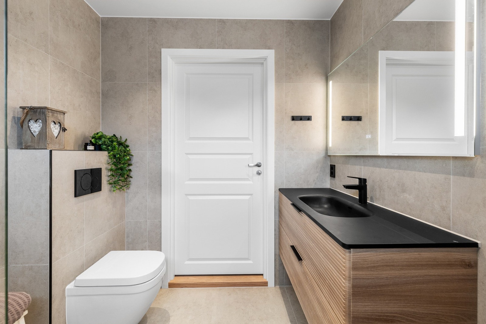 Stilfull baderomsinnredning med heldekkende sort servant og to romslige skuffeseksjoner, speil med integrert belysning samt vegghengt toalett. Galleribilde