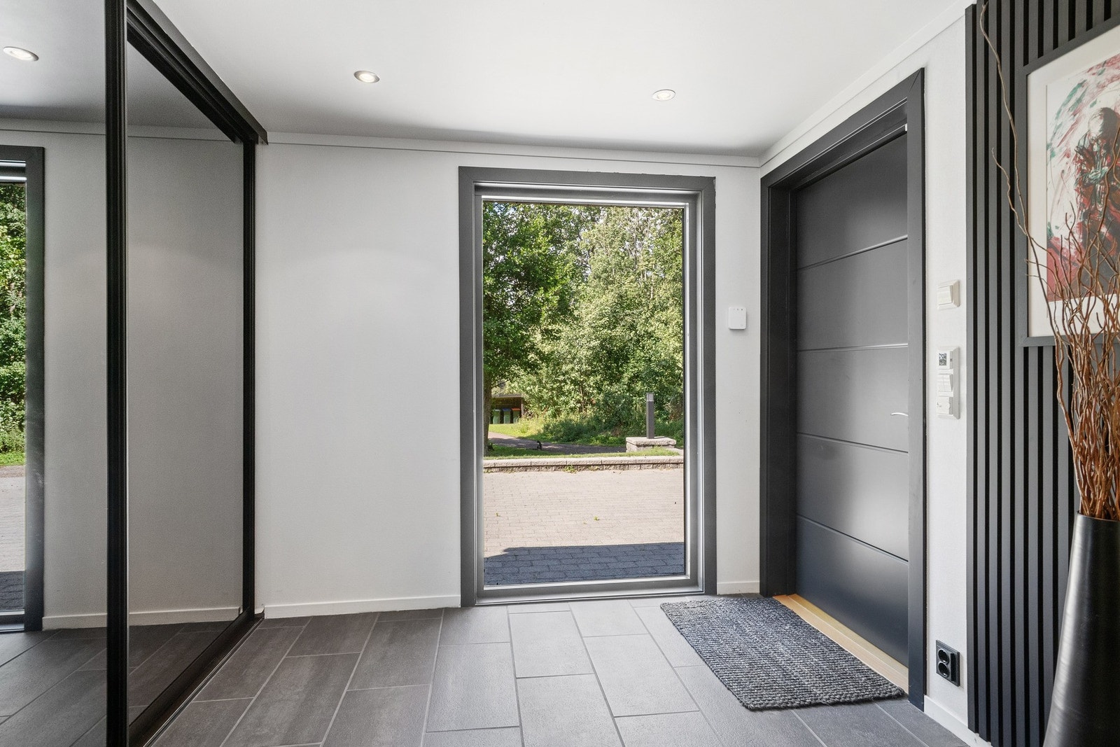 Romslig entré med hvitmalte, glatte vegger, grå fliser på gulvet og downlights i himlingen. Galleribilde