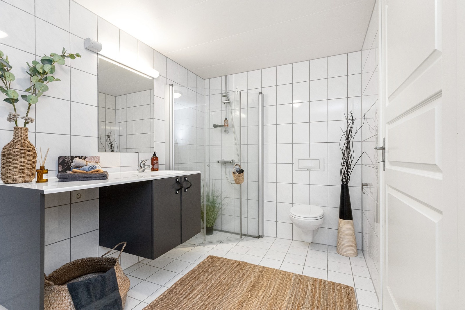 Hovedbadet er romslig og innbydende, med lyse fliser på vegg og gulv, dusjhjørne med innfellbare glassdører, vegghengt toalett og heldekkende servant med underskap Galleribilde