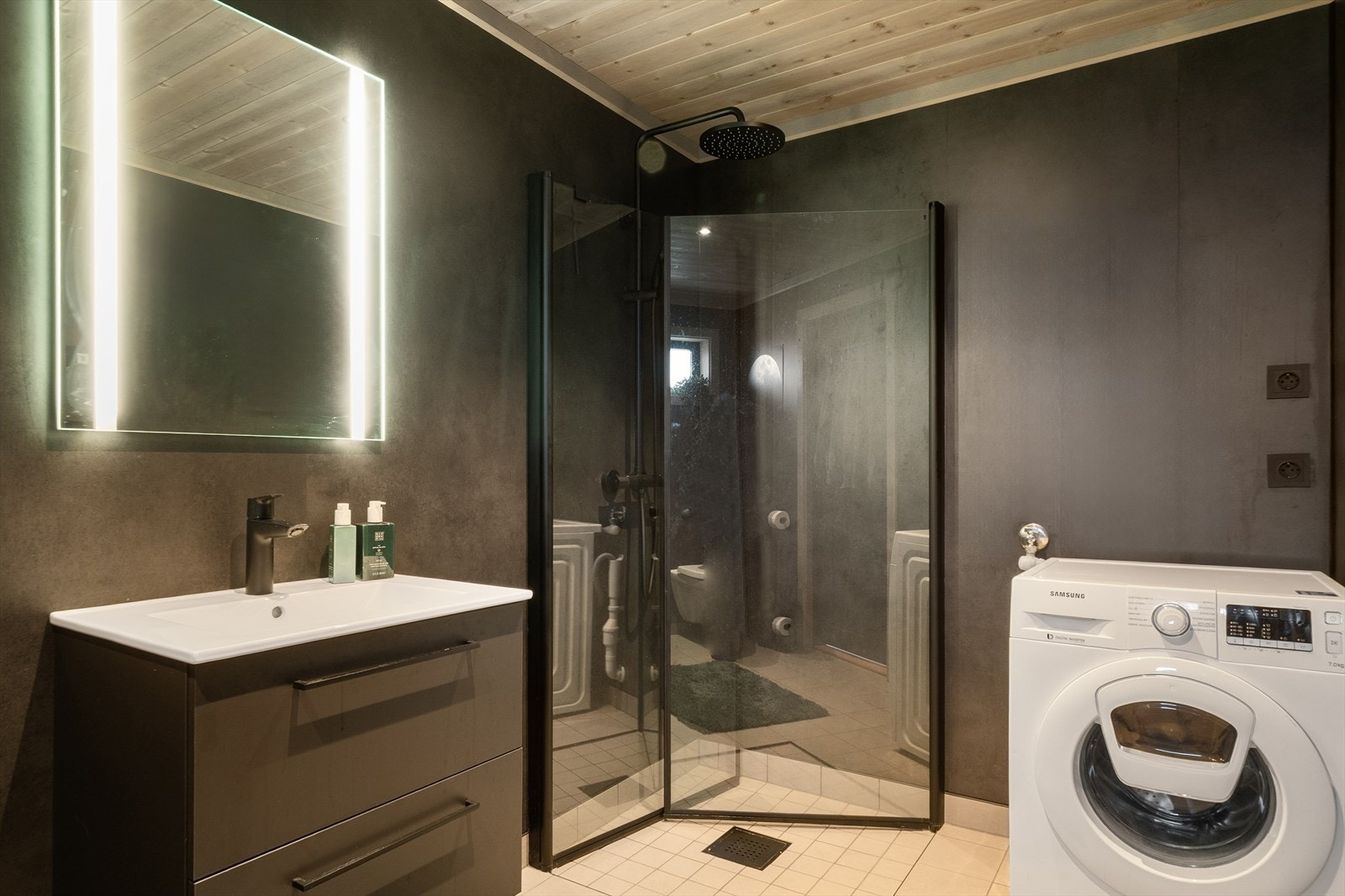 Badet har vegger med sokkelflis og baderomsplater, baderomsinnredning med heldekkende servantplate og ettgreps blandebatteri, dusjdører i glass, veggmontert termostatbatteri med dusjgarnityr, toalett med innebygget sisterne og varmekabler i gulv. Galleribilde