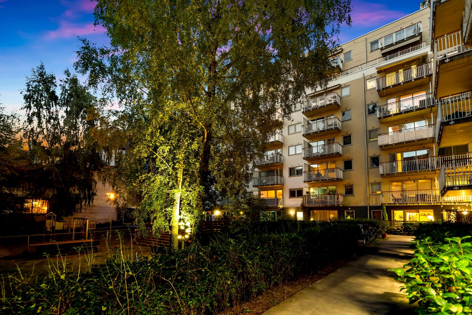 Kveldsbelysningen gir gårdsrommet en lun og trygg atmosfære, samtidig som den fremhever beplantningen og adkomsten til bygget. Galleribilde