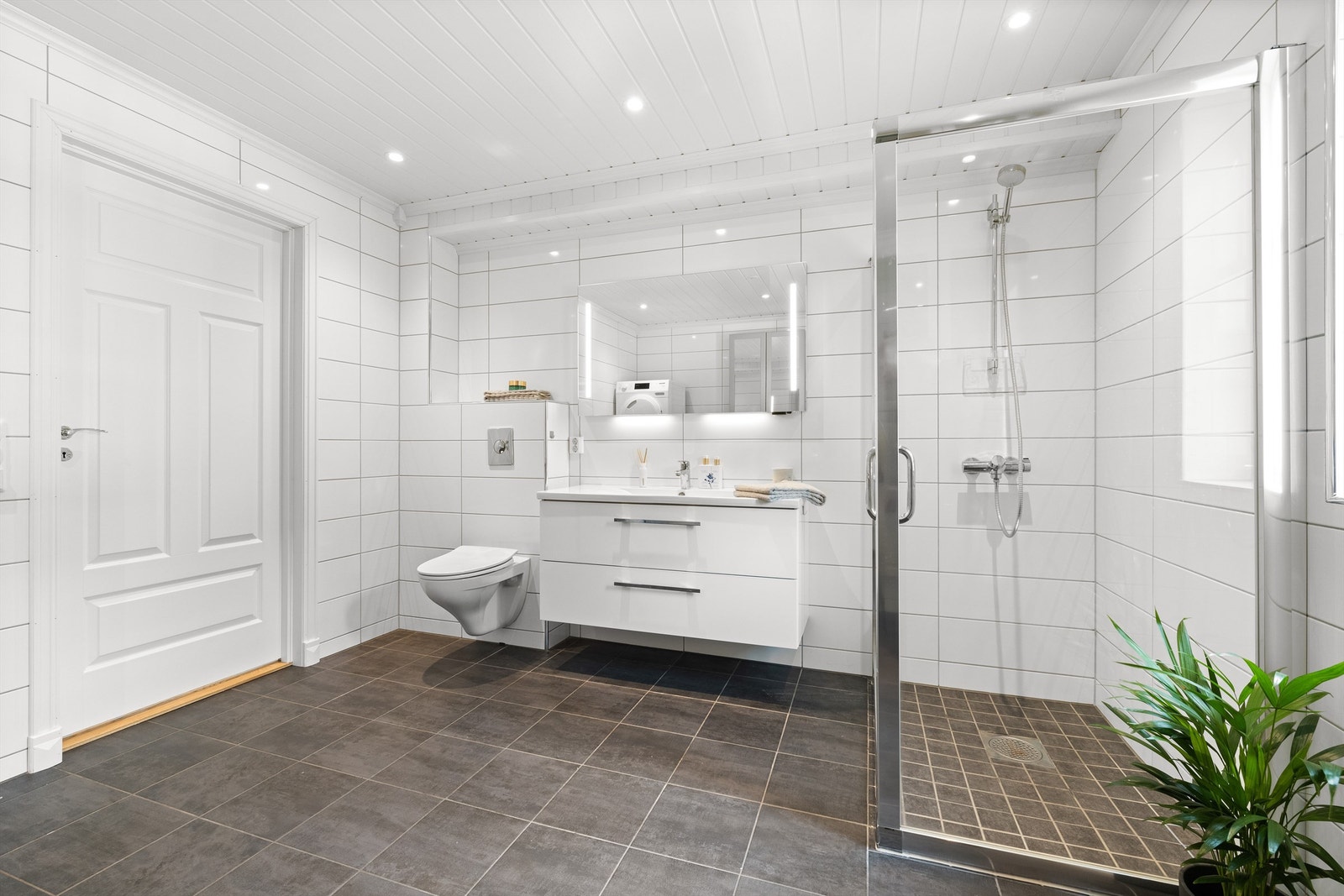 Baderom i 1. etasje er flislagt med varme i gulv. Høyskap og innredning med servant, speil med belysning samt dobbel stikkontakt på vegg, vegghengt toalett og dusjhjørne med skyvedør Galleribilde