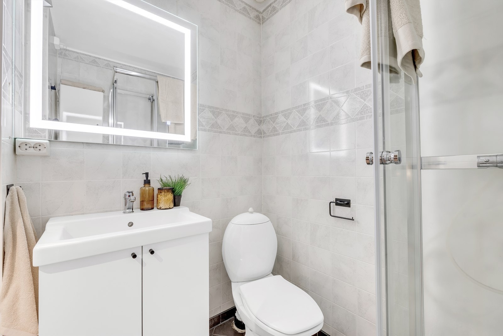 Badet inneholder dusjkabinett, gulvmontert toalett, innredning med servant og speil med lys. Galleribilde