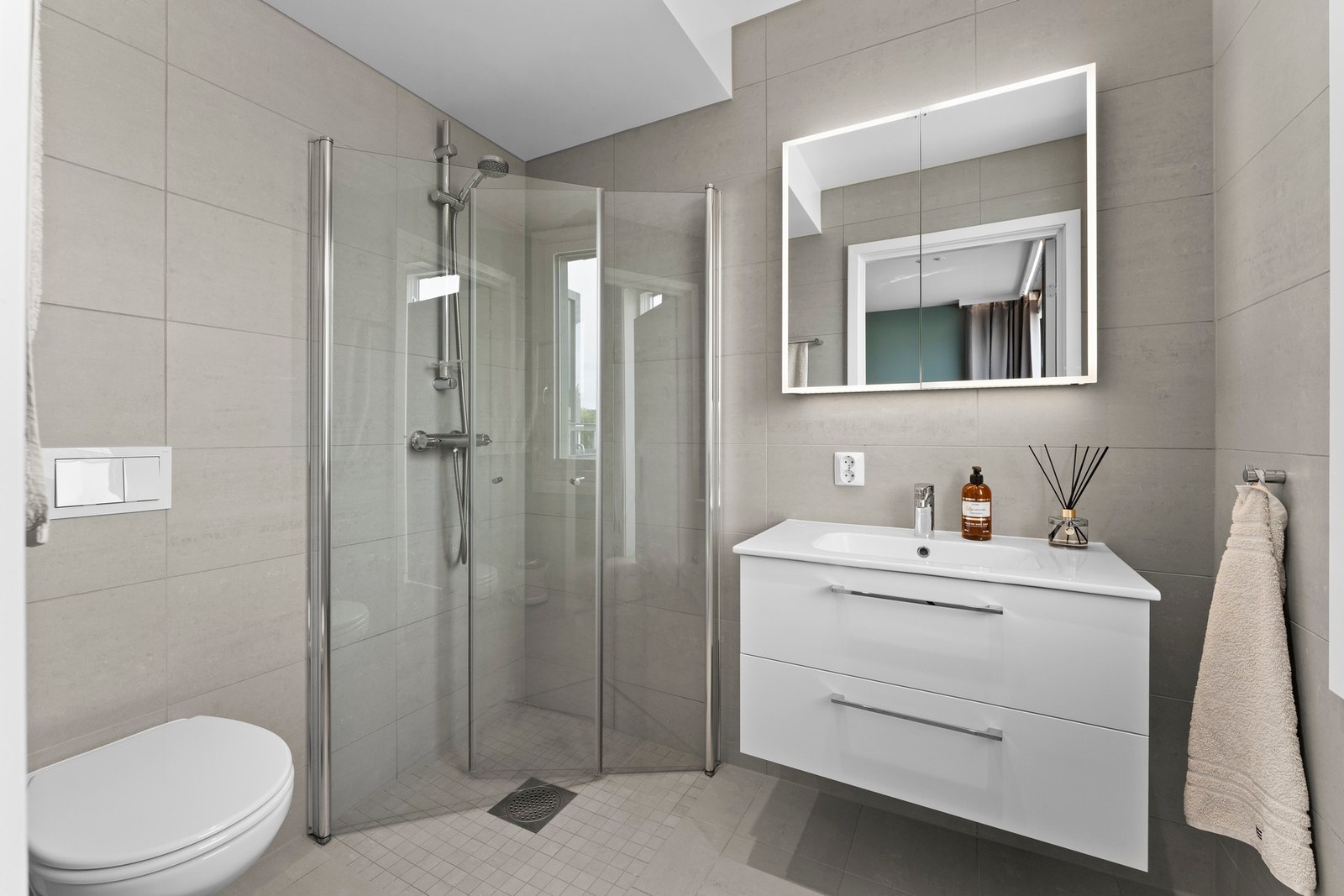 Badet er innredet med servantinnredning, speil med lys i dører på overskap på vegg over servant, vegghengt toalett og dusjhjørne med svingbare glassdører. Galleribilde