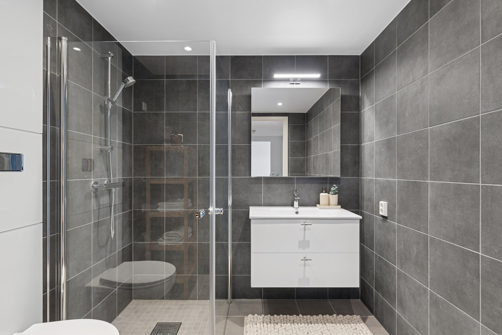 Videre er rommet utstyrt med baderomsinnredning med servant og skuffer, speil, dusjhjørne med glassdører og vegghengt toalett. Det er opplegg for vaskemaskin og gulvvarme i gulvet. Galleribilde