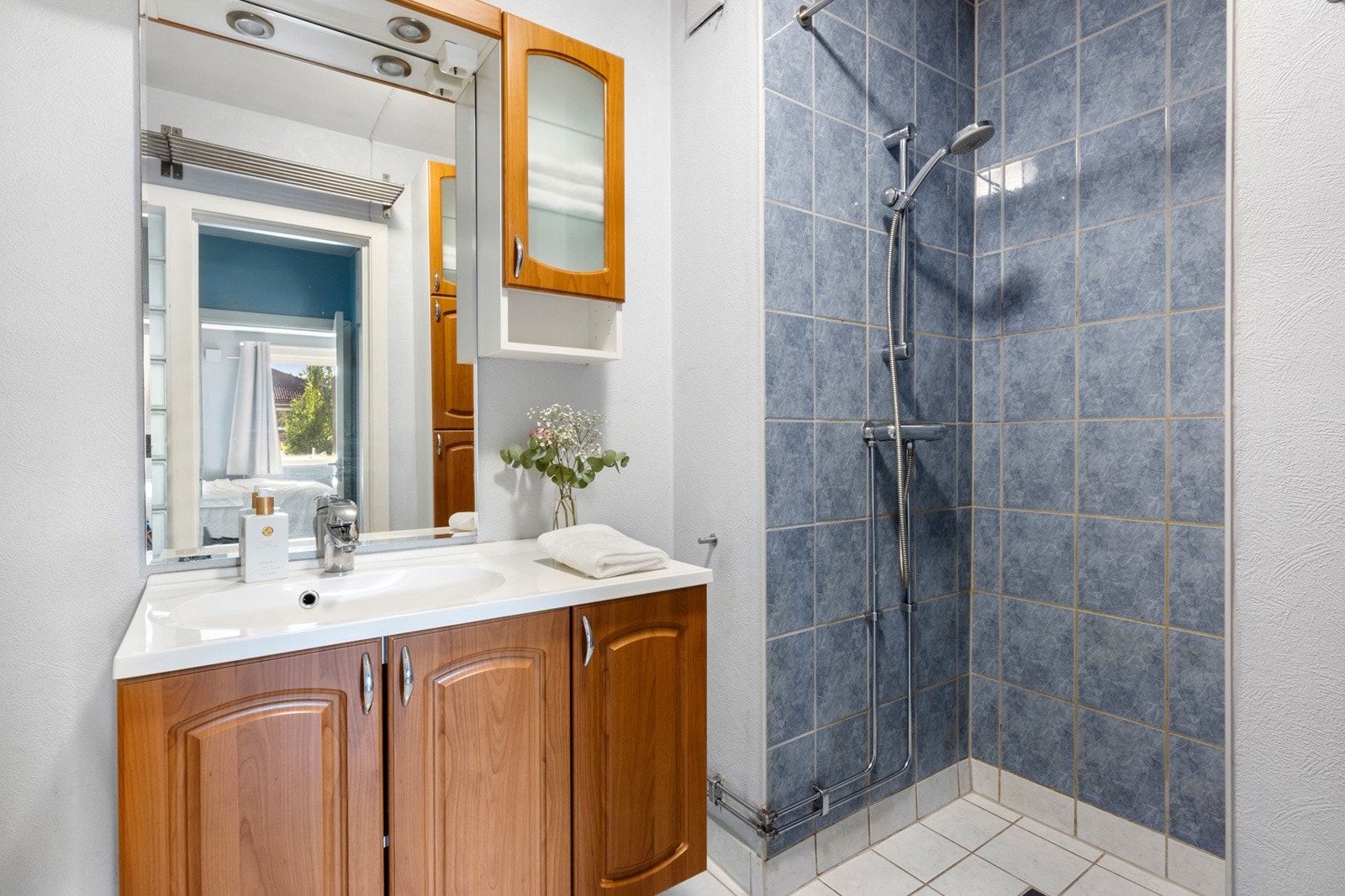 Flislagt bad med vegghengt servantinnredning med skapdører og ovenpåliggende servant med ett-greps armatur, speil med overlys, dusjhjørne og toalett. Galleribilde