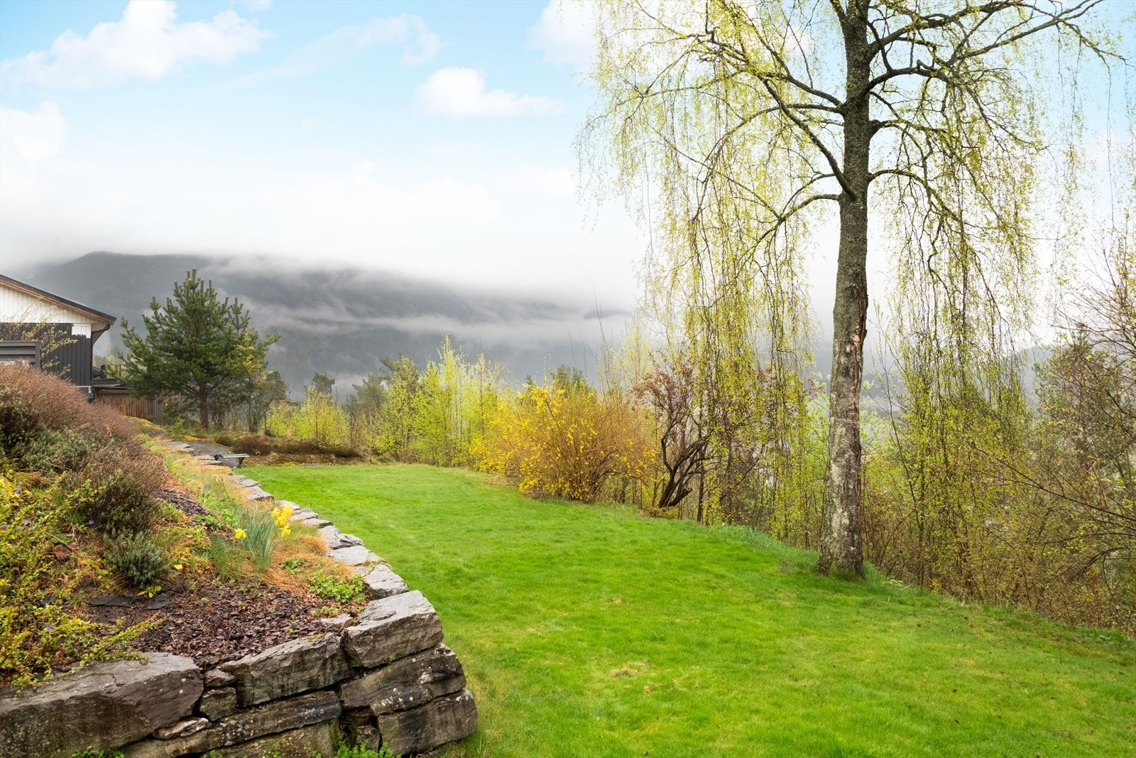 Mellom øvre og nedre nivå av hagen mot sør er det etablert en støttemur av naturstein. Hagen gir fin plass til lek og aktivitet for store og små. Galleribilde
