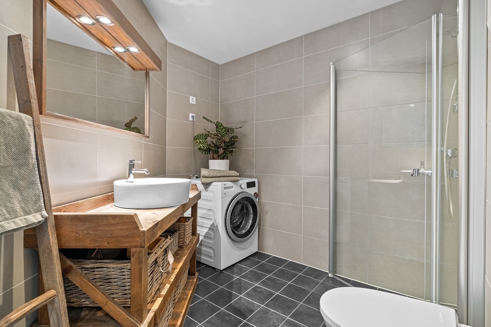 Badet er lekkert helfliset med varme i gulvet. Det inneholder en praktisk dusjnisje med innfellbare dusjdører, vegghengt toalett, opplegg for vaskemaskin/tørketrommel, samt en stilig baderomsinnredning med tilhørende speil og belysning ovenfor. Galleribilde