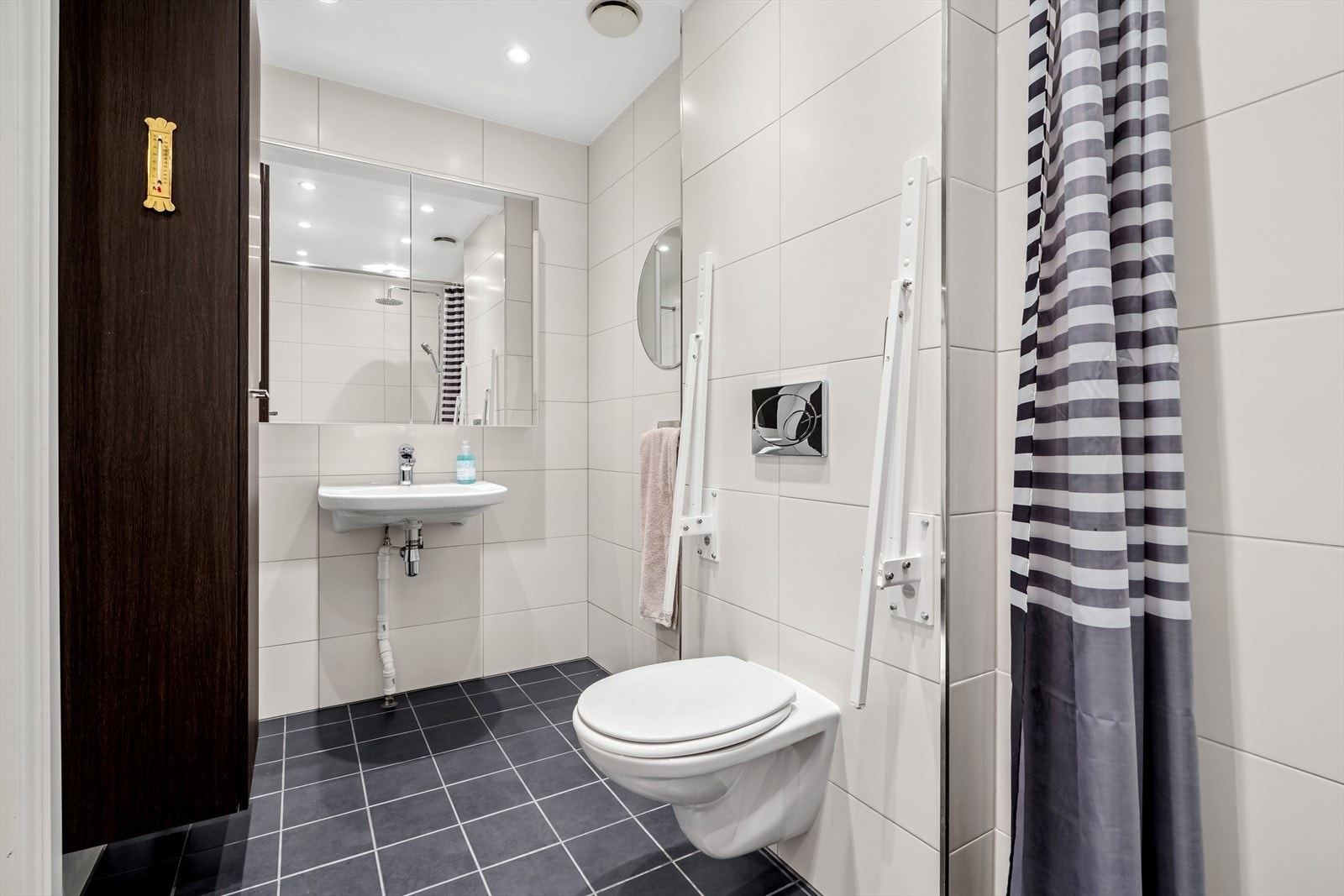 Badet er flislagt med gulvvarme. Rommet har vegghengt servant, høyskap, veggmontert toalett m/håndtak og dusjhjørne. Galleribilde