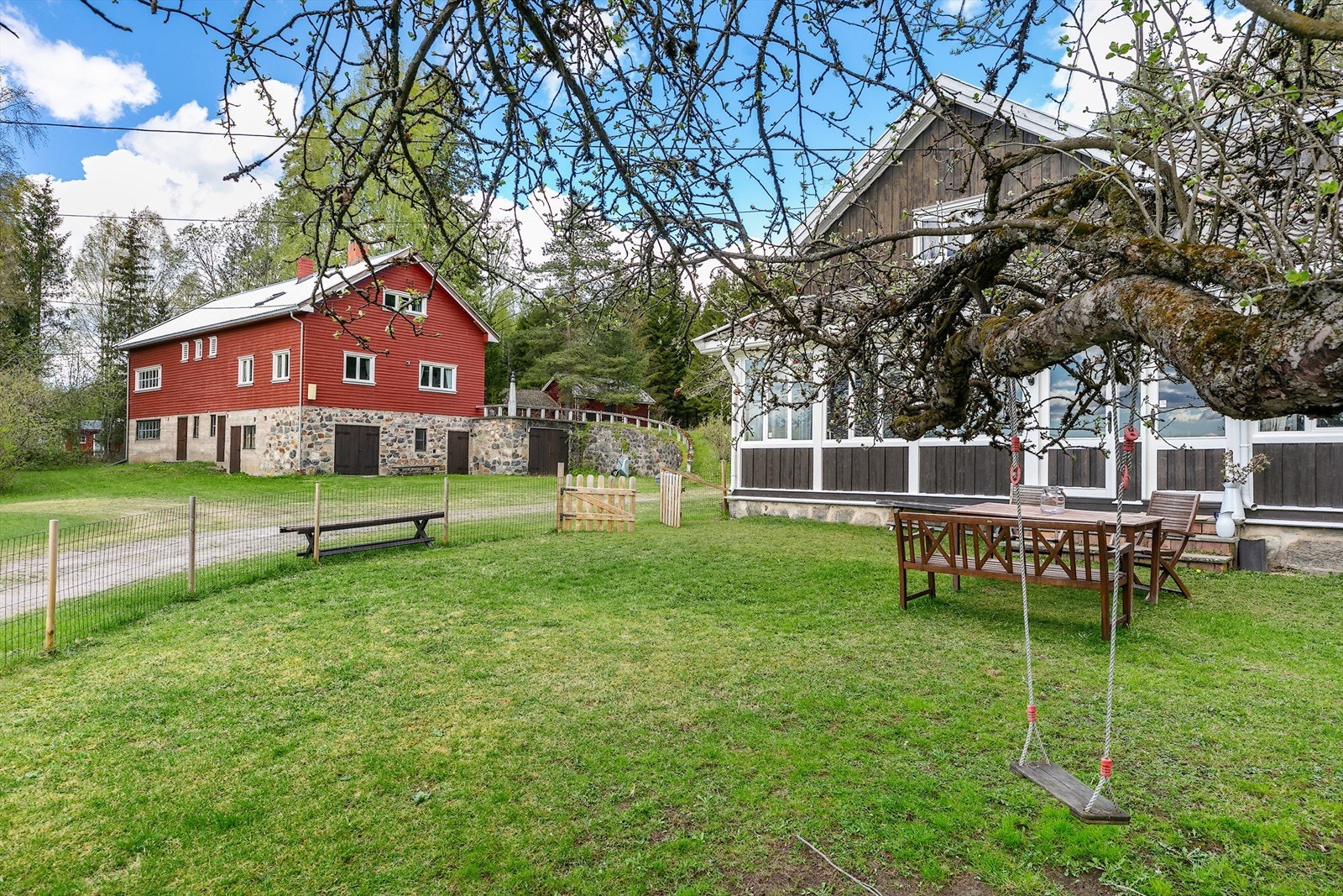 Utsyn mot eiendommens to bygg - den sjarmerende rødmalte låven med natursteinsmur, og det koselige hovedhuset med innglasset veranda. Galleribilde