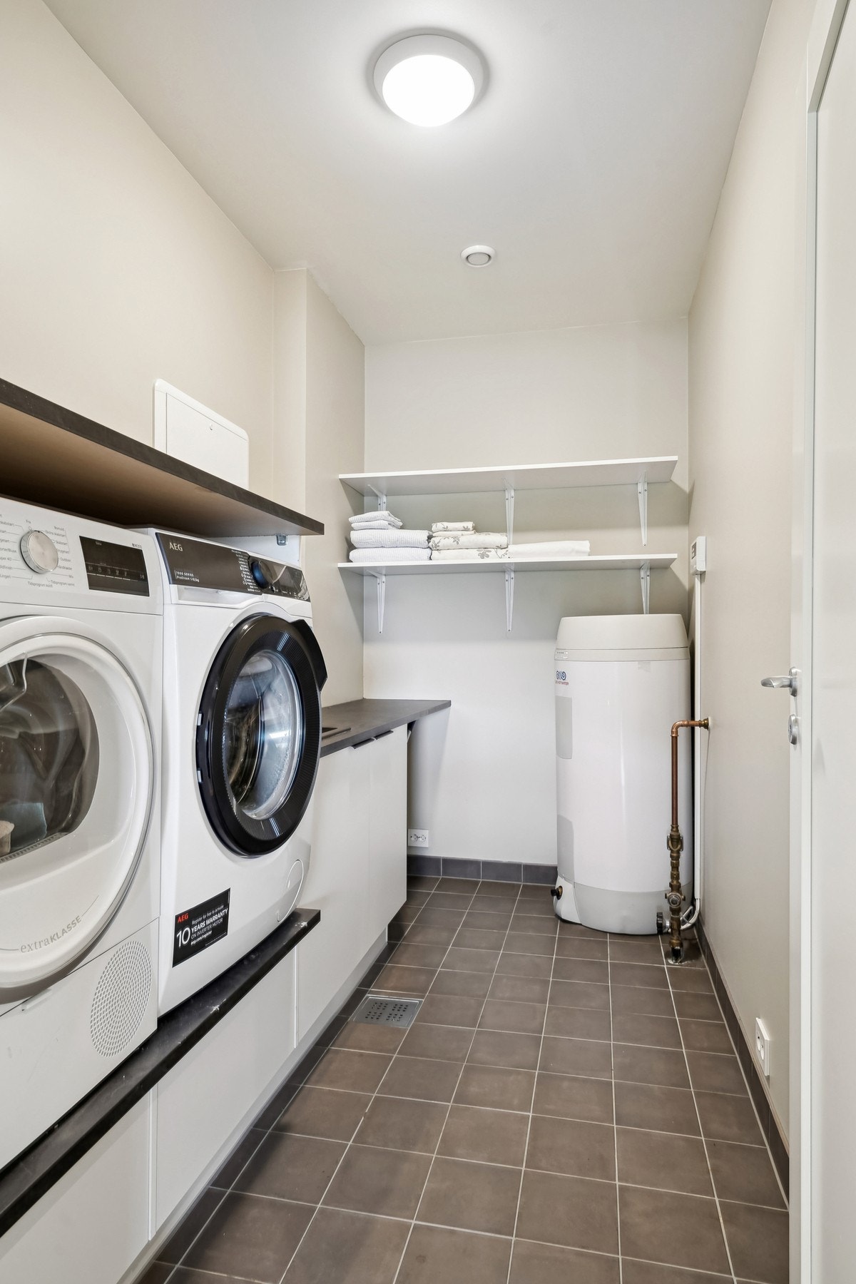 Praktisk vaskerom med flislagt og varmekabler. Innredet med hyller, innredning med utslagsvask, opplegg for vaskemaskin og tørketrommel. Galleribilde