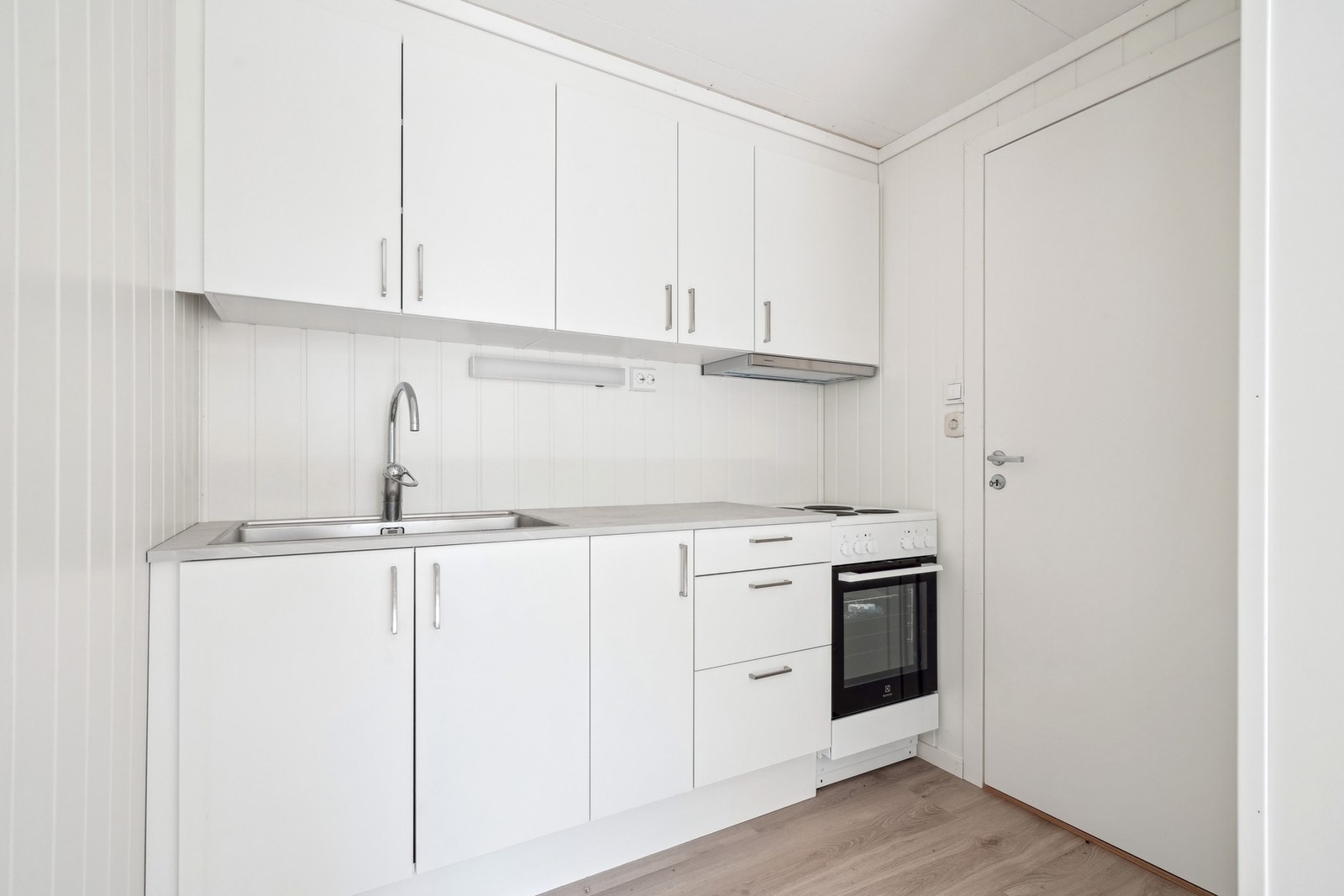 Kjøkkeninnredningen har hvite glatte fronter og en laminat benkeplate. Det er kjøkkenventilator med avtrekk ut over komfyren. Galleribilde