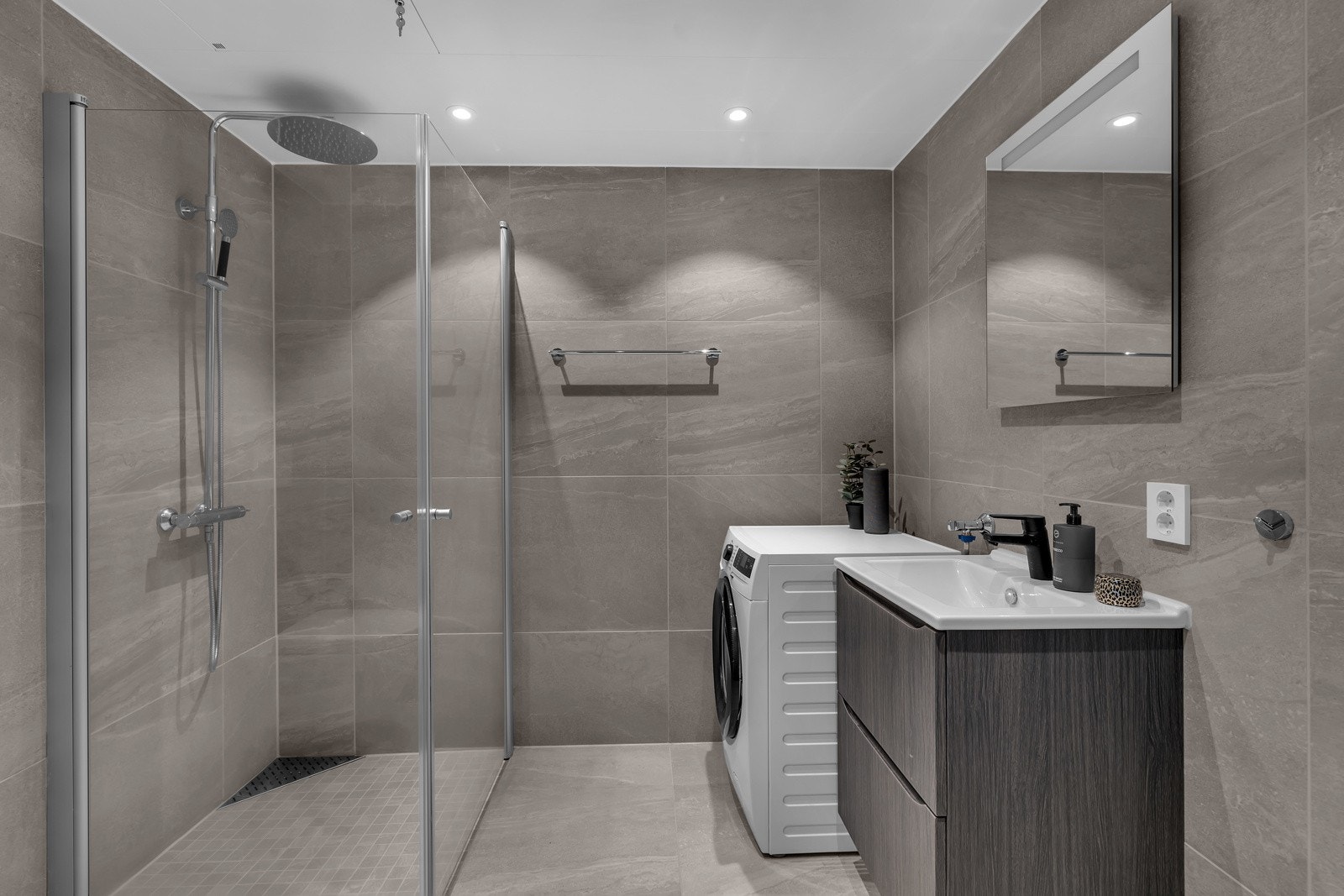 Badet er pent flislagt på gulv og vegger, har innfelt belysning i tak, innfellbare glassdører og opplegg for vaskemaskin og tørketrommel Galleribilde