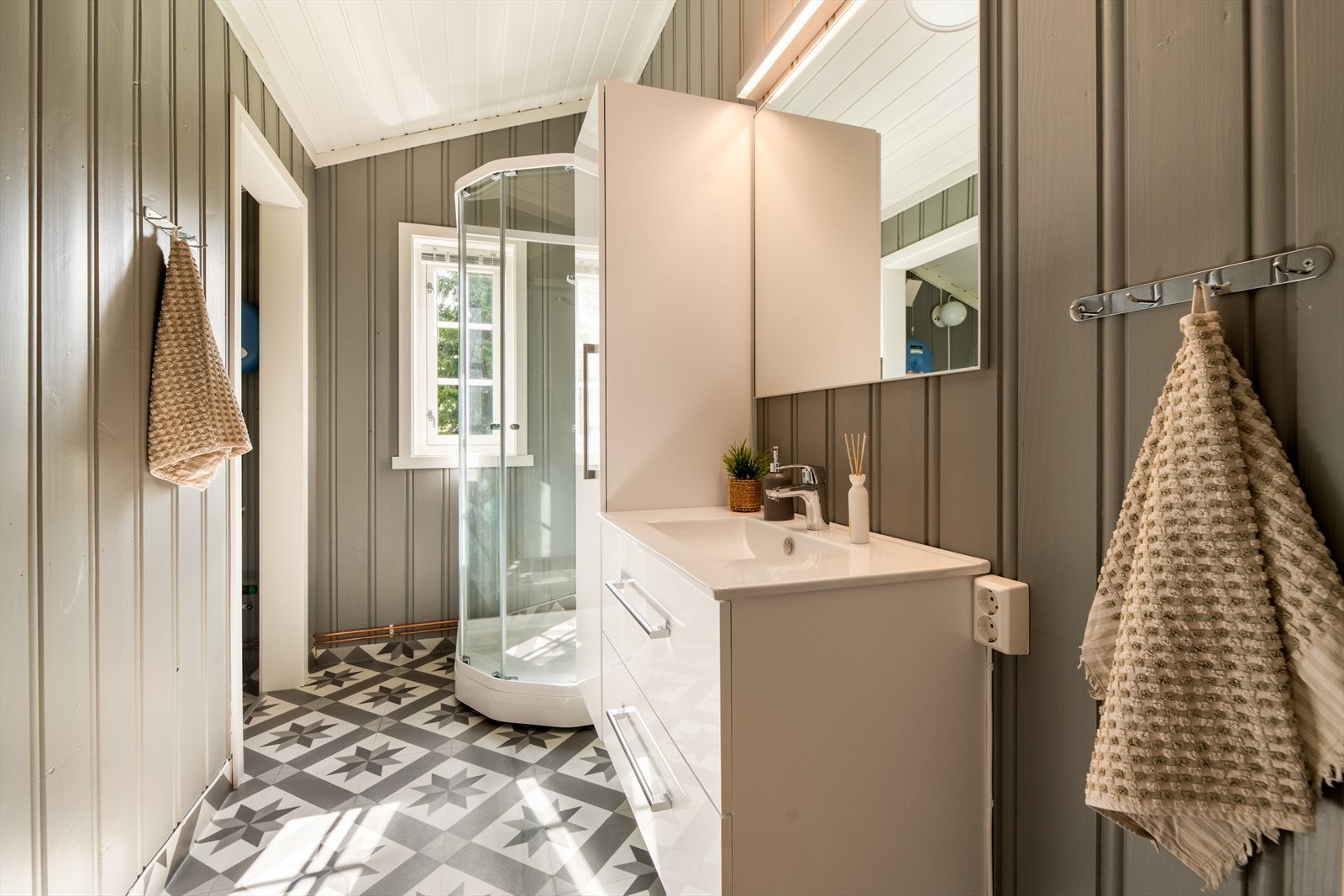 Pent bad fra 2020 med gulvbelegg og trepanel på vegger. Her er det dusjkabinett og innredning med heldekkende hvit vask, underskuffer med slette fronter og speil. Det er i tillegg et høyskap med ekstra oppbevaringsplass. Rommet har naturlig ventilering. Galleribilde