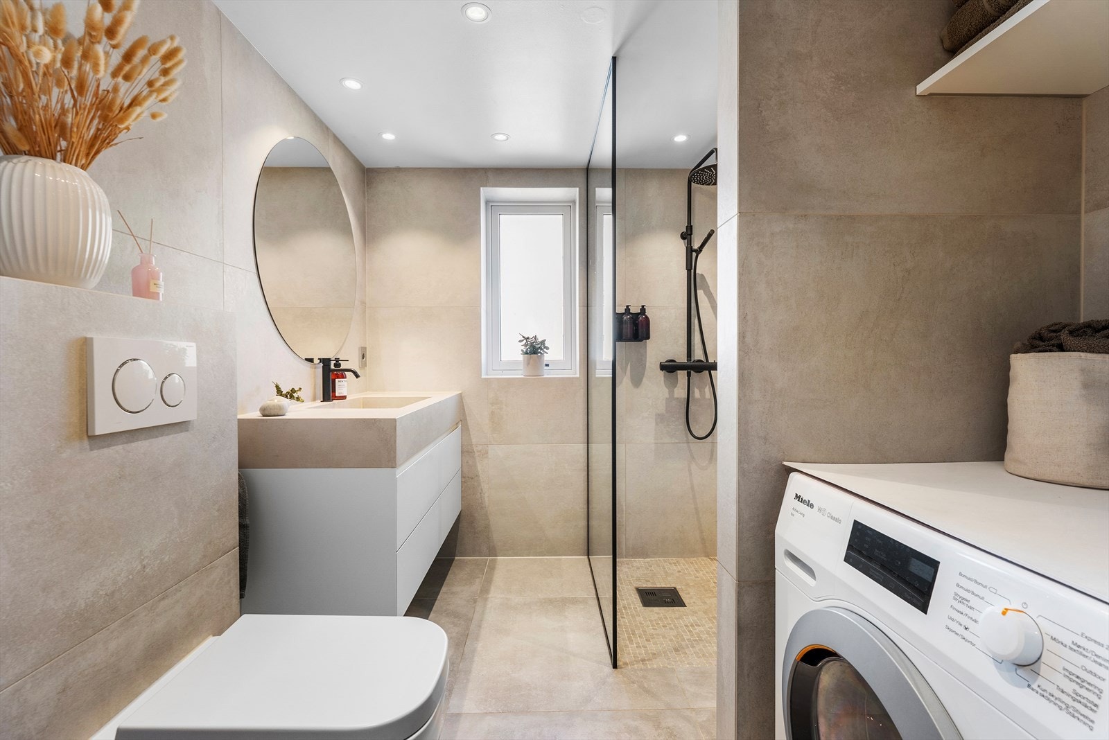 Lekkert og flislagt bad med varmekabler, samt stilren innredning og overflater fra 2020 - Praktiske hyller over opplegg for vaskemaskin. Arbeidet er gjort av Bella Fliser og Design Galleribilde