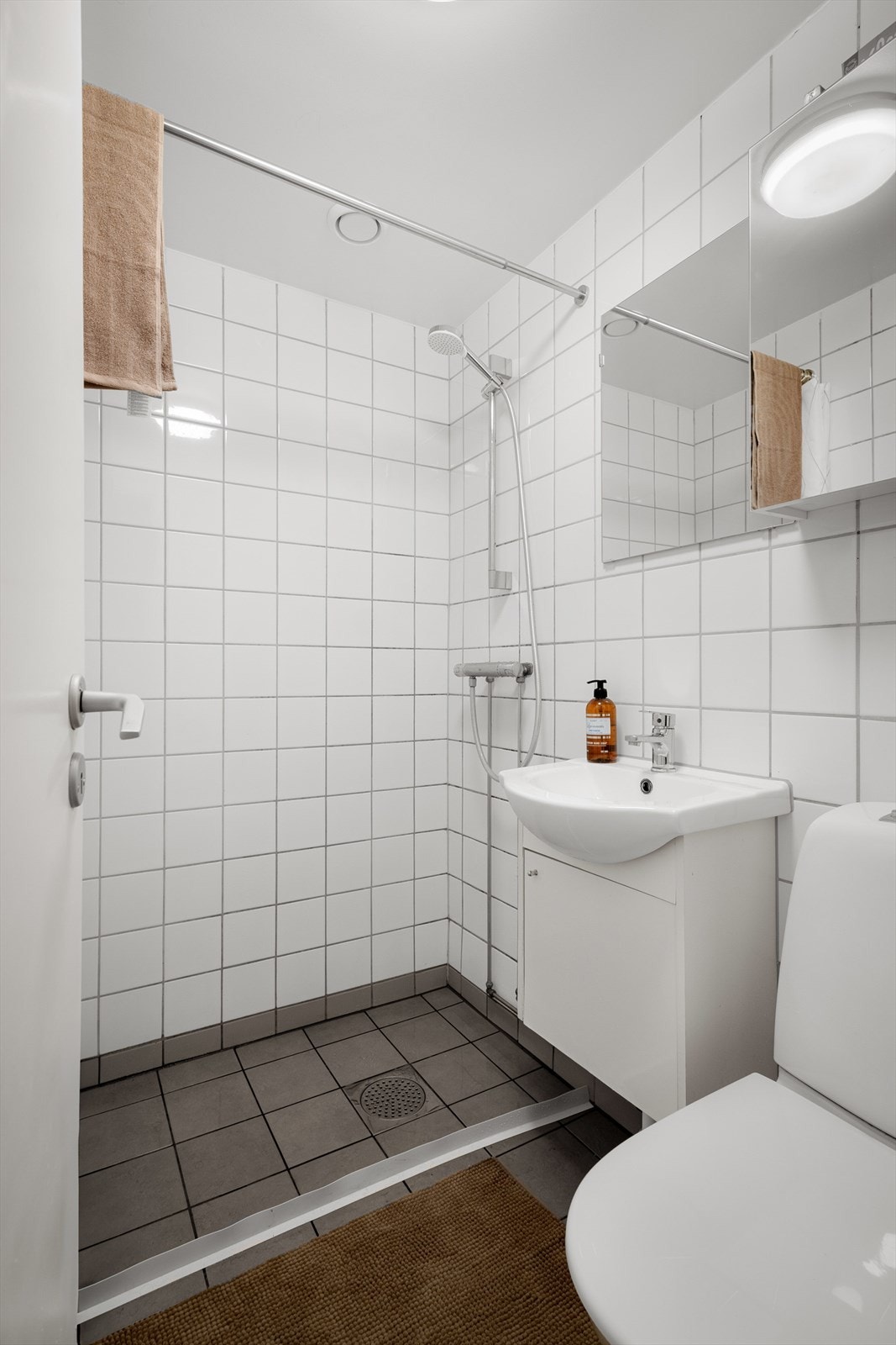 Servantskap med glatte fronter og heldekkende servant, speil på vegg over servant, gulvmontert klosett, dusjhjørne med forheng, sluk og termostatstyrt blandebatteri. Galleribilde