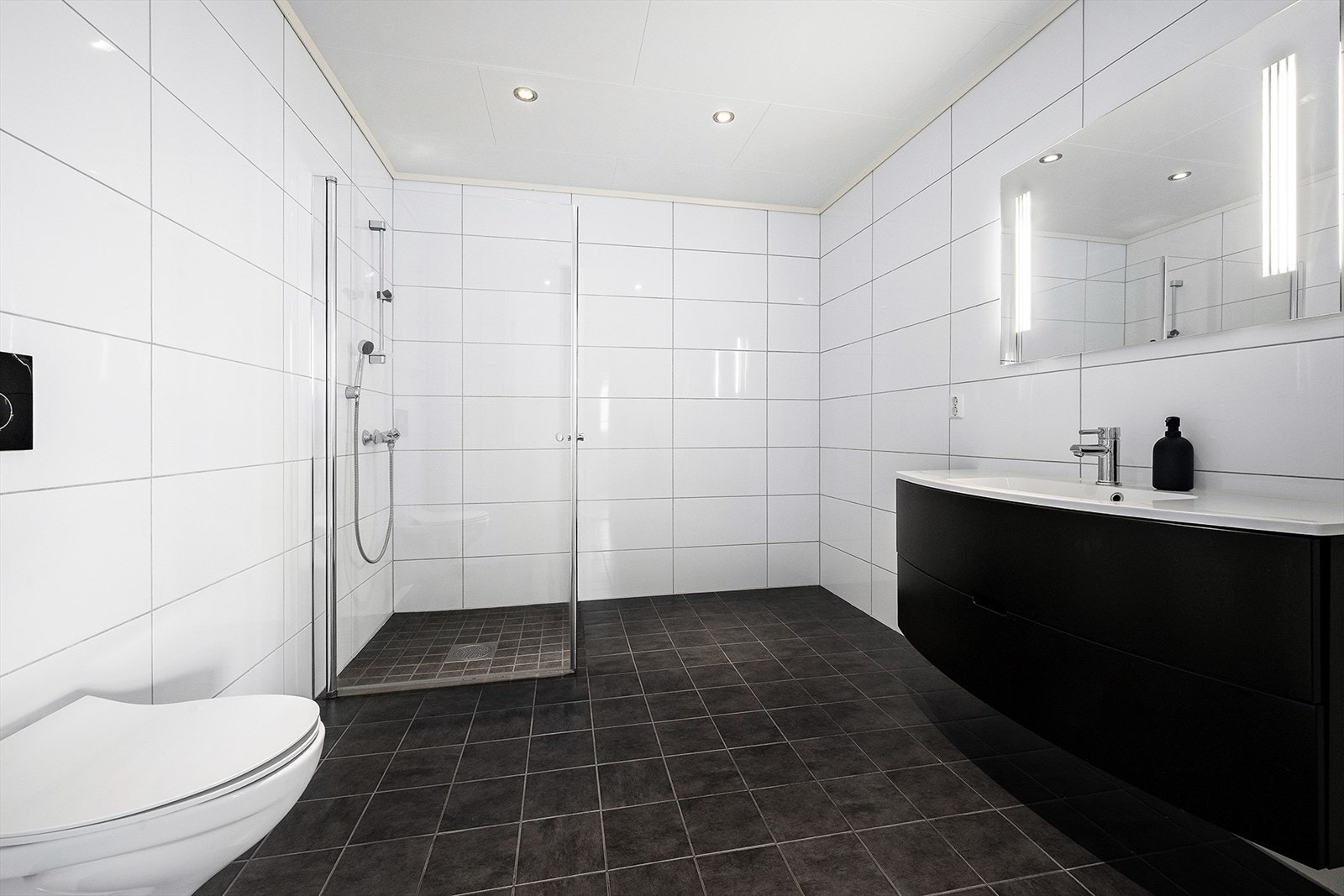 Badet er meget romslig og er utstyrt med dusjhjørne, veggfestet WC og baderomsinnredning med servant og speil. Galleribilde