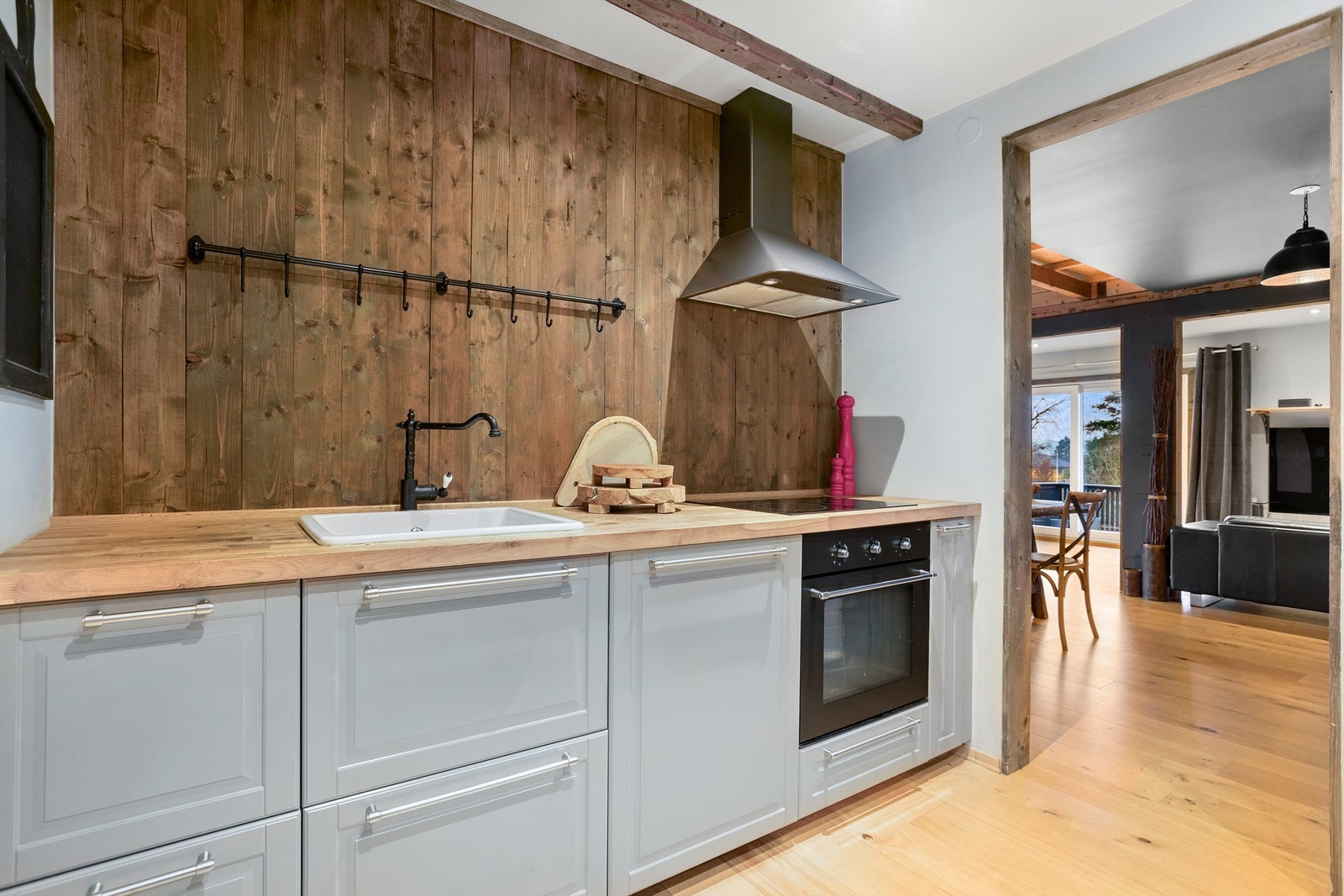 Benkeplater i tre og stein, stekeovn med keramisk platetopp og vegghengt ventilator. Vannrør av rør-i-rør, synlige avløpsrør av plast og nedsenket oppvaskkum med ett-greps armatur Galleribilde