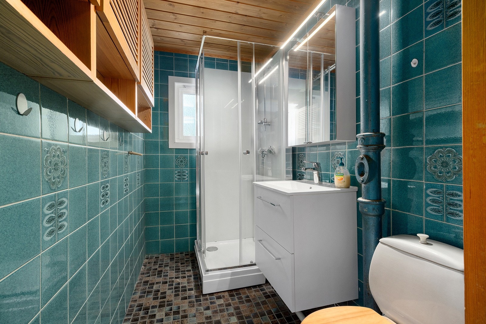 Eldre bad med moderniseringsbehov. Mosaikkfliser på gulv, flislagte vegger og malte himlingsflater. Termostatstyrt gulvvarme. Servantskap med glatte fronter og heldekkende servant, speilskap med lys, gulvmontert klosett og dusjkabinett med skyvedør. Galleribilde