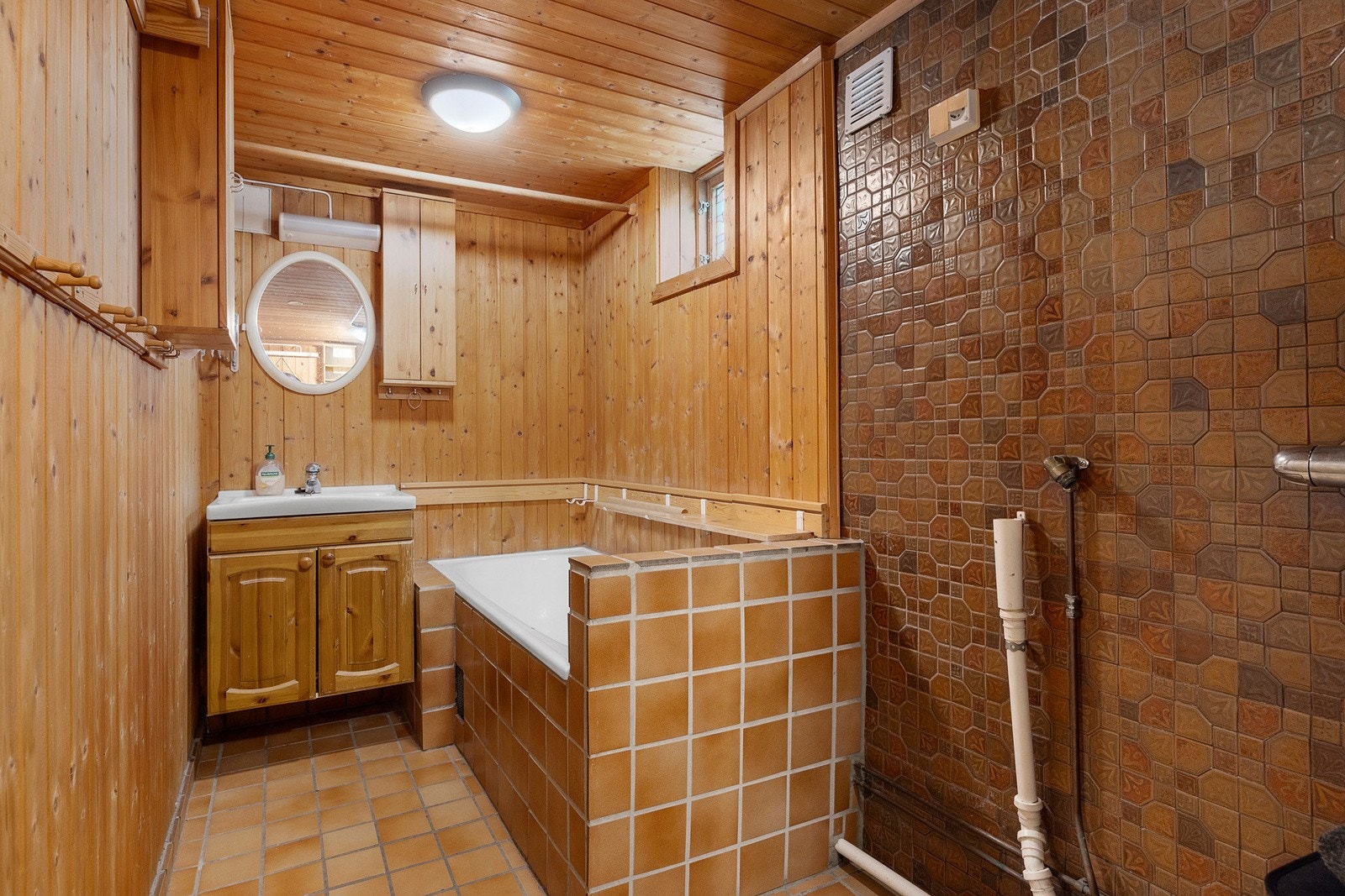 Eldre baderom med flis- og paneloverflater, dusjhjørne, badekar, servantskap og opplegg for vaskemaskin. Galleribilde