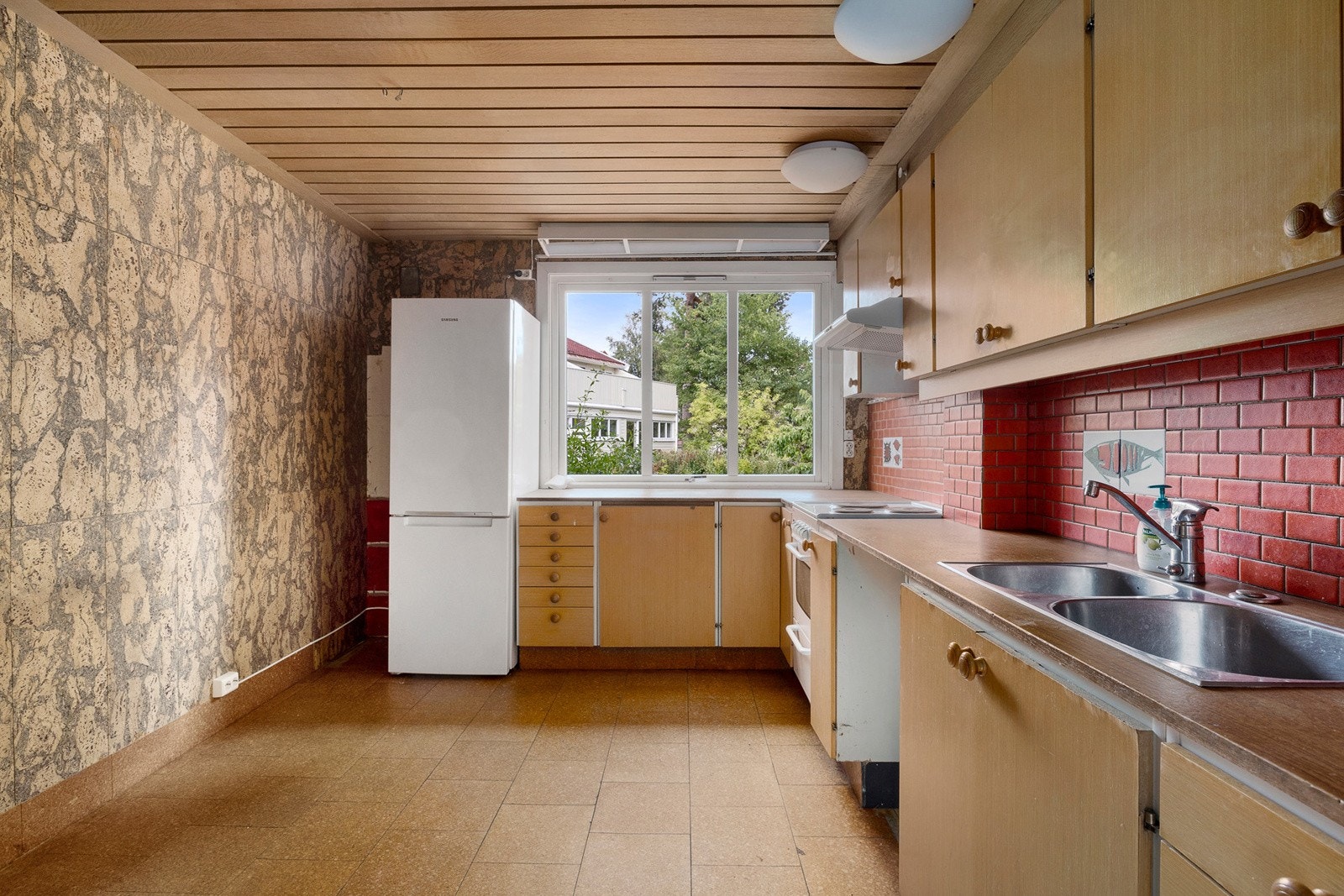 Eldre kjøkkeninnredning med glatte, finerte fronter, laminatbenkeplate med nedfelt kum i rustfritt stål, samt flisfelt på vegg over benk. Galleribilde