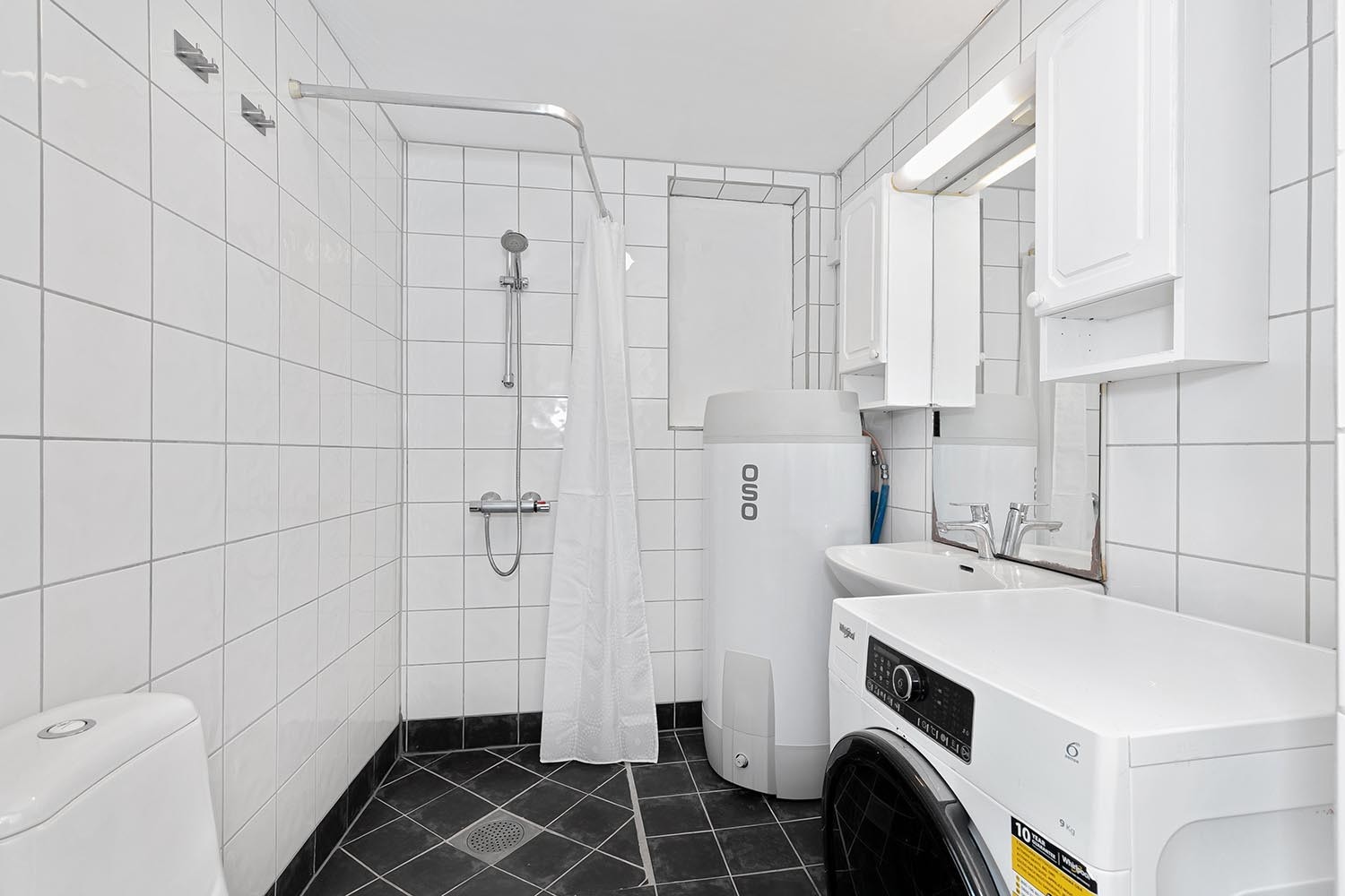 Pent flislagt bad med toalett, servant, vaskemaskin og hjørnedusj. Galleribilde