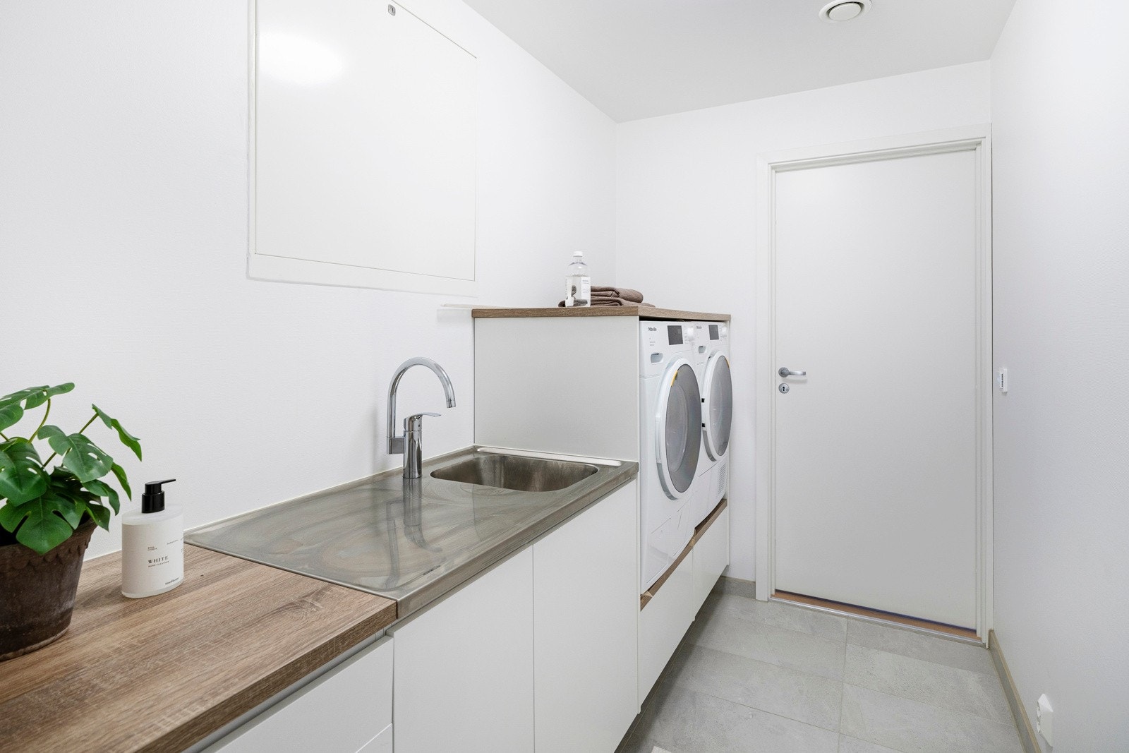 Tvers over gangen for bad 2 er det et pent og praktisk vaskerom med hvitmalte vegger og lysegrå fliser på gulv med vannbåren gulvvarme. Galleribilde