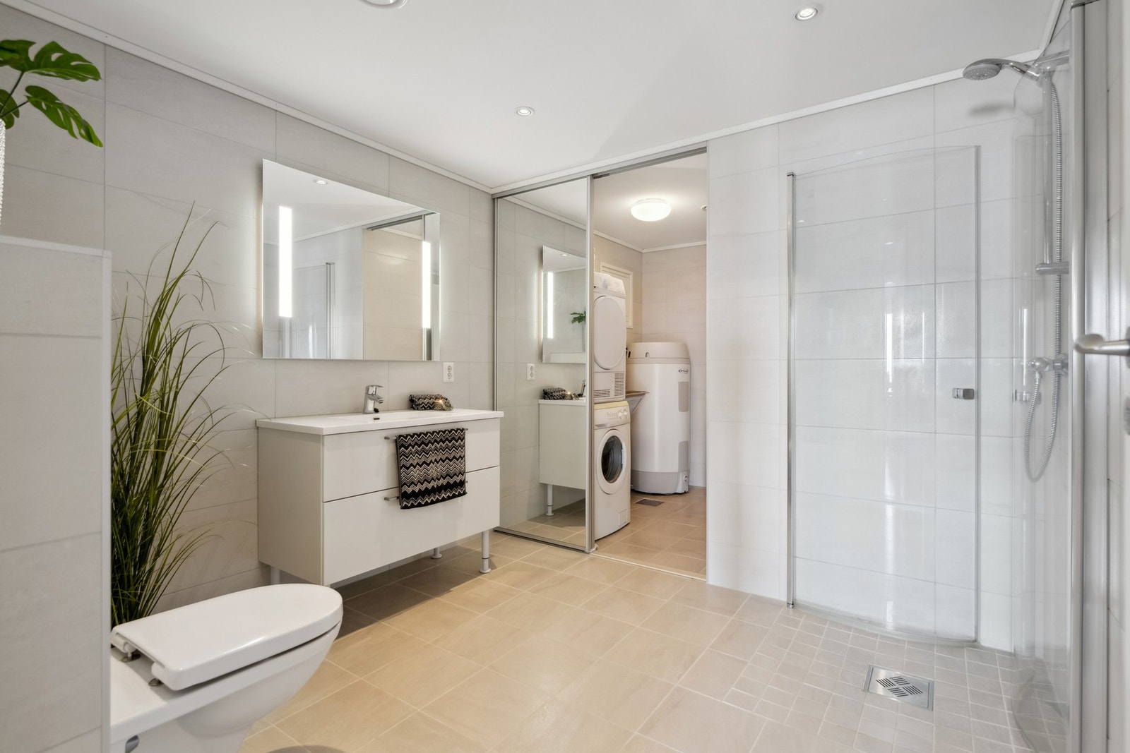 Flislagt bad/vaskerom med varme i gulvet. Badet er delt i to funksjonelle soner, hvor vaskeromsdelen er adskilt med skyvedører med speilfronter Galleribilde