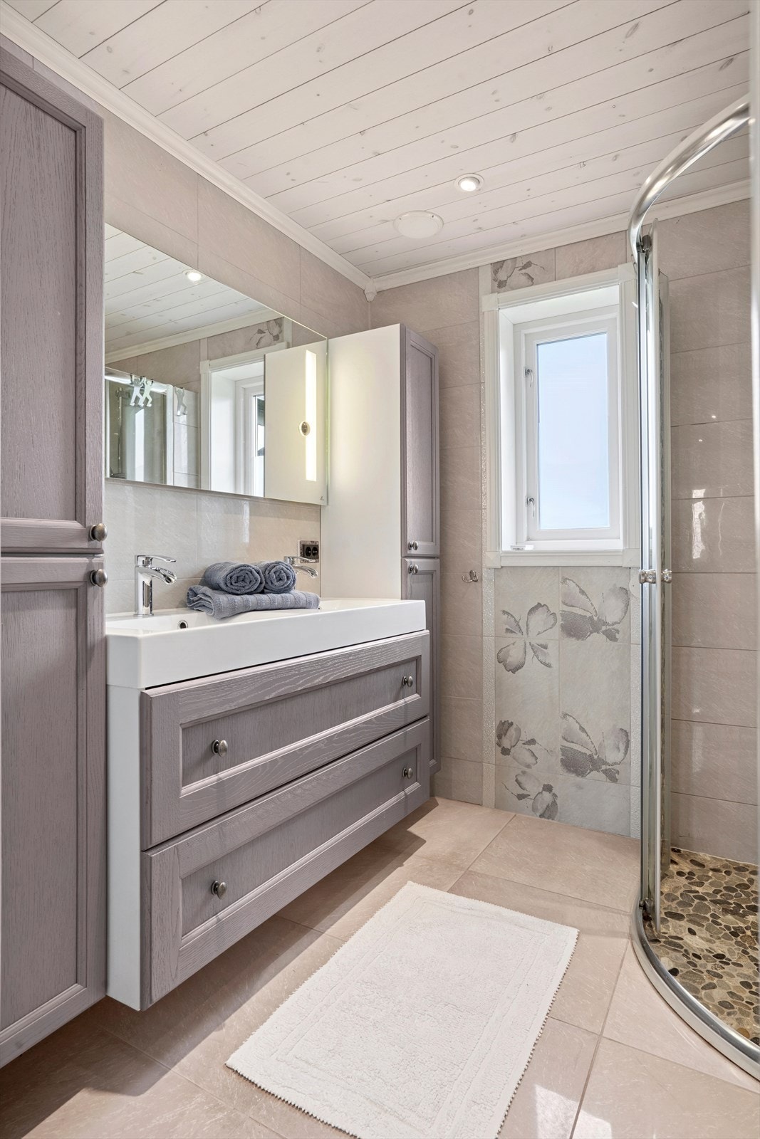 Badet består av speil med belysning, dobbelservant med oppbevaringsseksjon, dusjhjørne og veggmontert toalett. Galleribilde