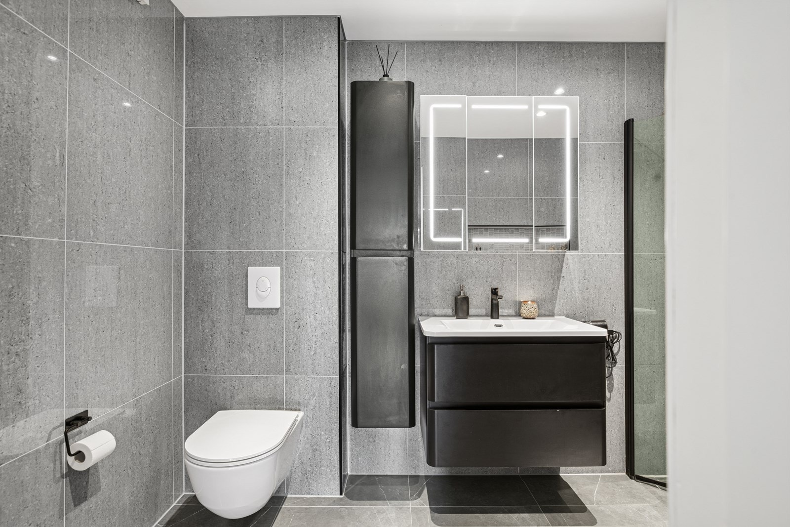 Badet er innredet med sort, glatt servantskap, vegghengt toalett med skjult sisterne og dusjhjørne med glassvegger. Downlights i himlingen gir god og jevn belysning, og mekanisk avtrekk sørger for effektiv ventilasjon. Galleribilde