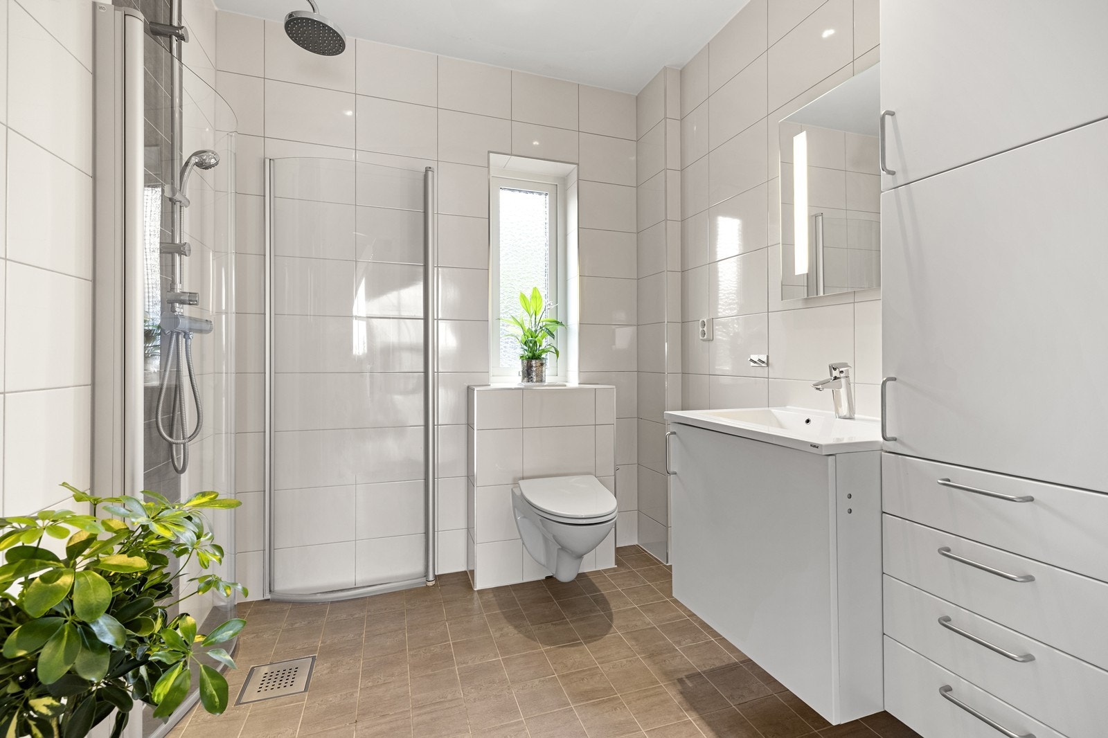 Baderom med flislagte gulv og vegger. Badet innehar vegghengt toalett, servant med skap og dusjnisje med innfellbare glassdører. Galleribilde