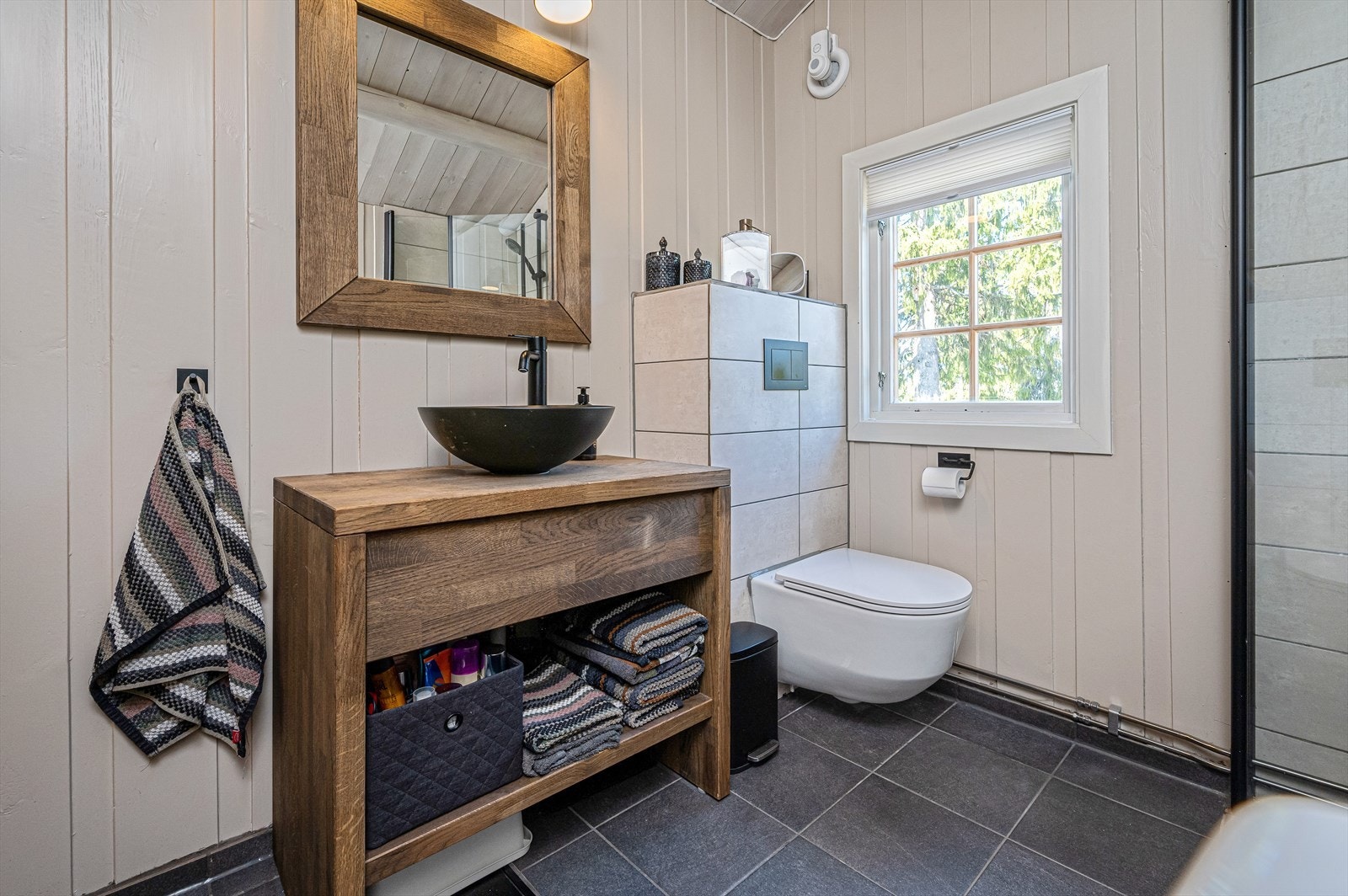 Badet består av en egendesignet baderomsinnredning med servant og speil over, vegghengt toalett, dusjdører og opplegg til vaskemaskin. Galleribilde