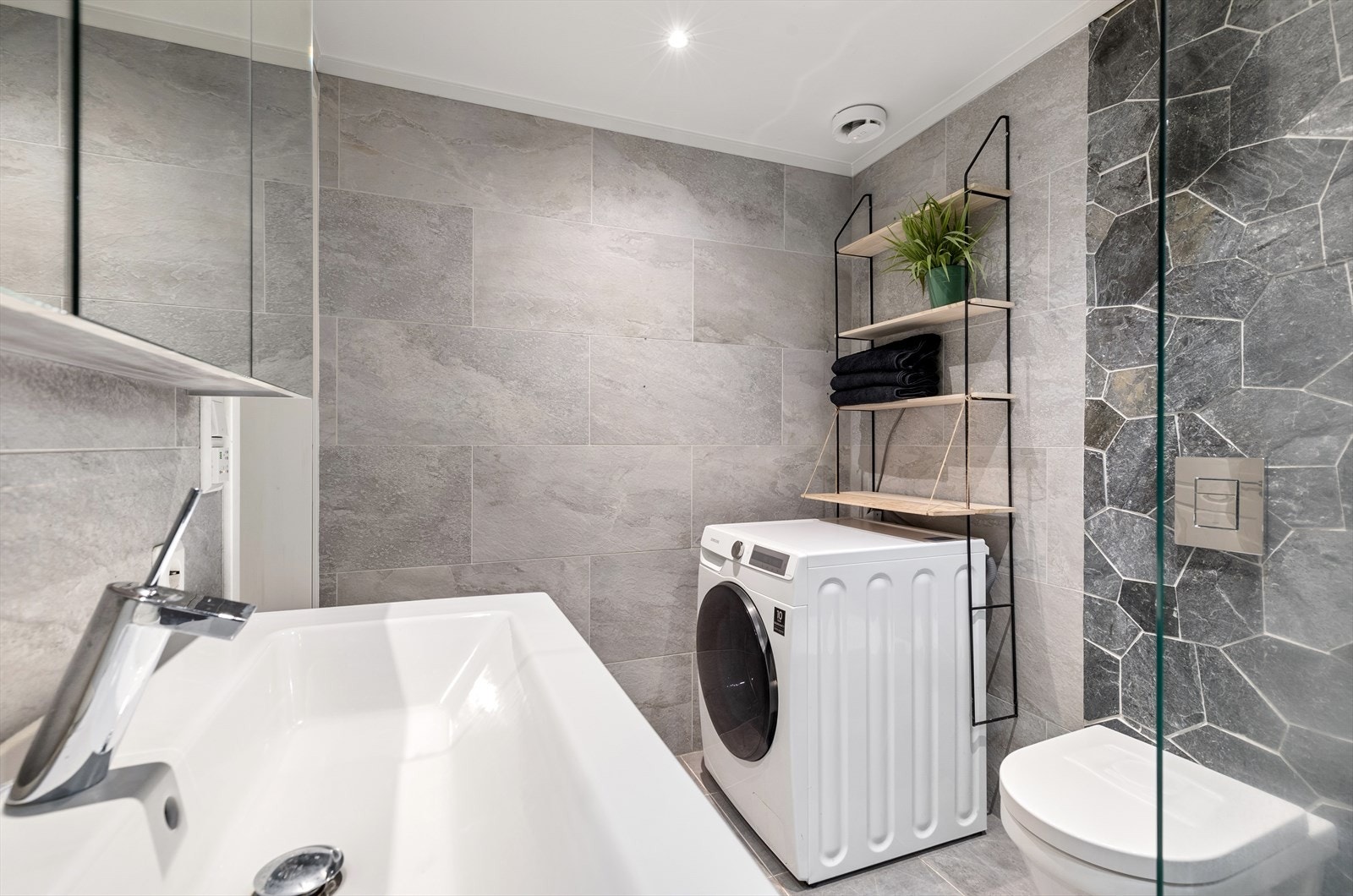 Meget lekkert bad med store fliser på gulv og vegger, samt en kontrastvegg bak toalett og bak dusjoppheng Galleribilde