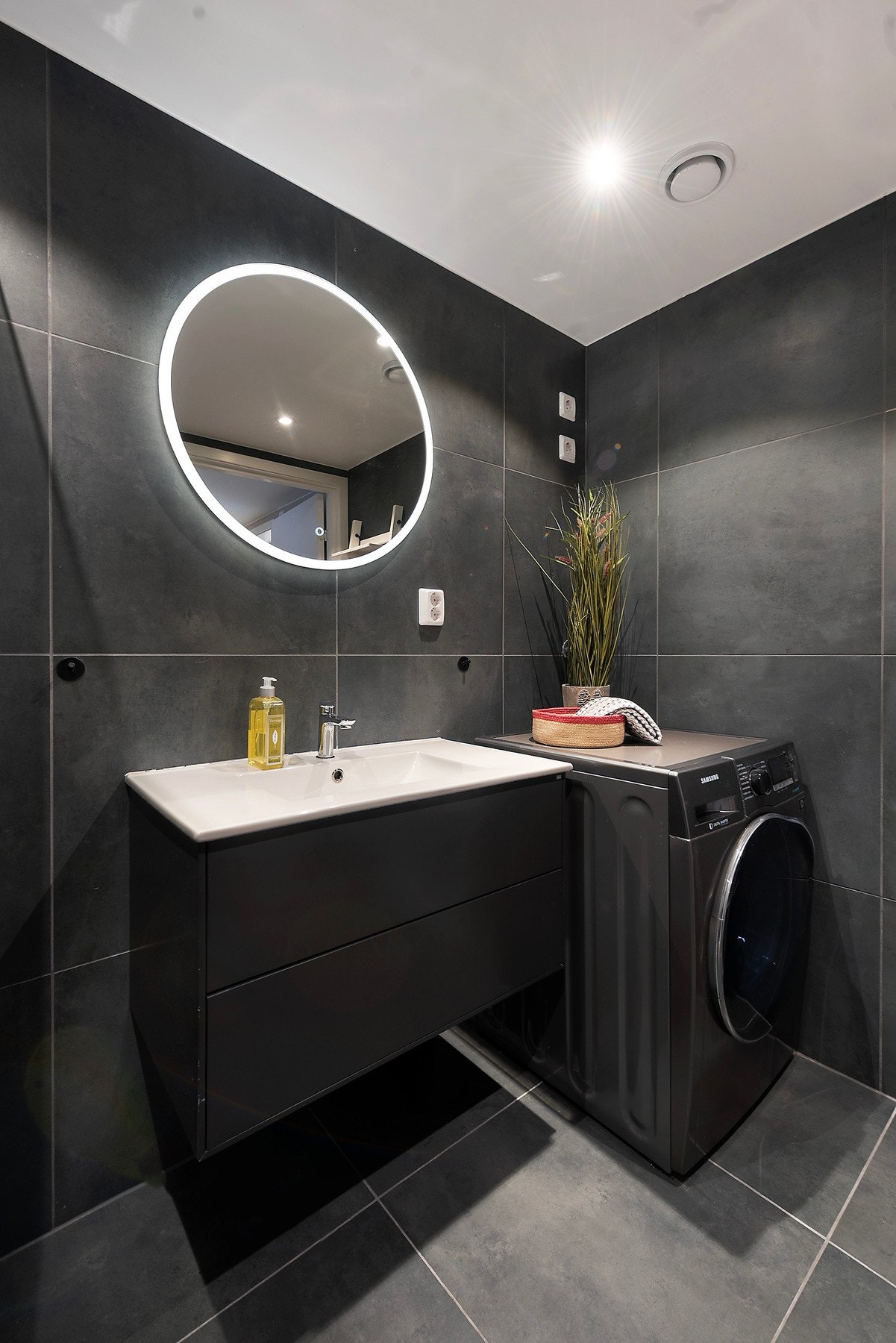 Innredningen består av en heldekkende servant på sort skuffeseksjon, rundt speil med rammebelysning, et veggmontert toalett, og et dusjhjørne med innfellbare glassdører Galleribilde