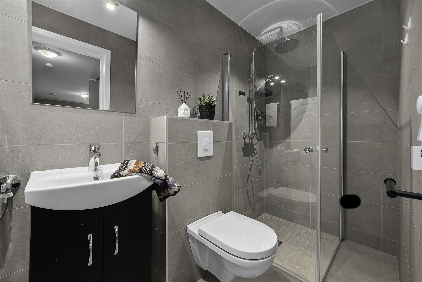 Bad/vaskerom med fliser på gulv og vegger, servant med skuffer, dusjnisje, opplegg for vaskemaskin og vegghengt toalett. Galleribilde