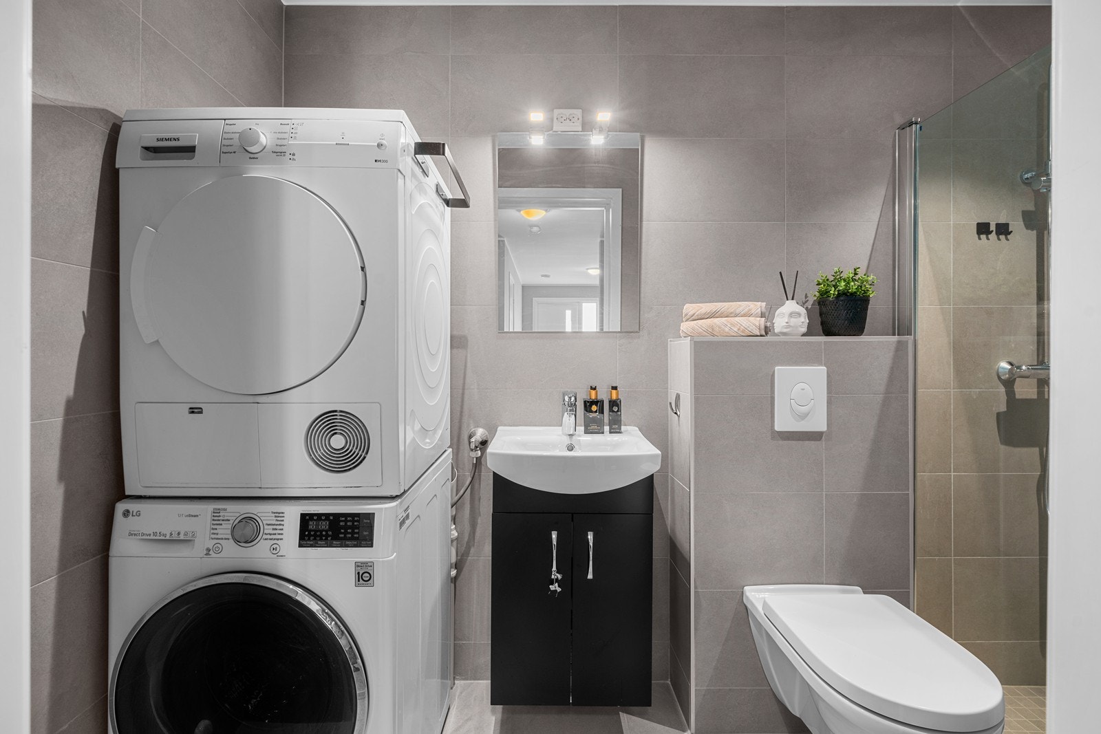 Bad/vaskerom med fliser på gulv og vegger, servant med skuffer, dusjnisje, opplegg for vaskemaskin og vegghengt toalett. Galleribilde