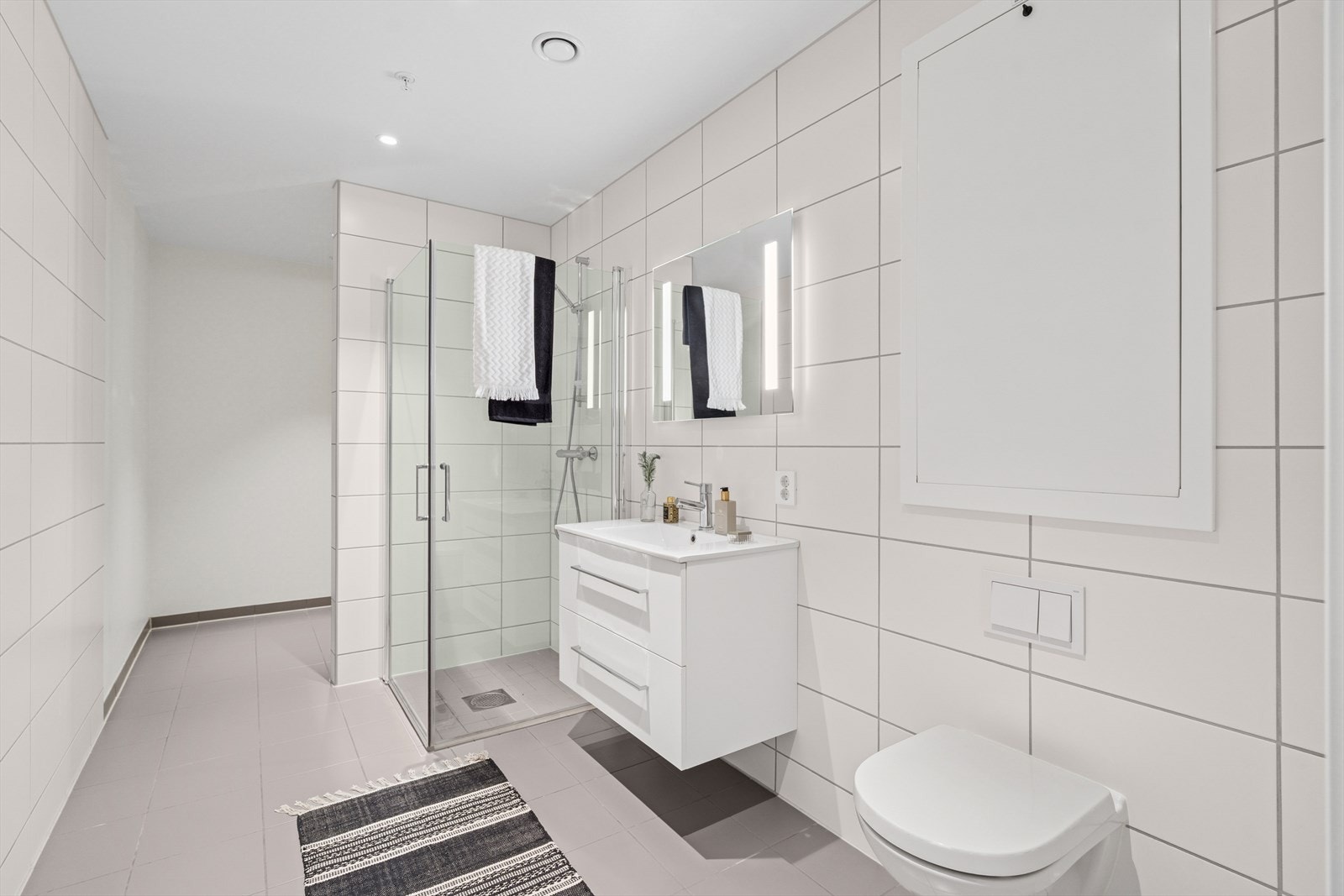 Badet har en innredning med servant, veggmontert toalett og et dusjhjørne i en nedsenket dusjnisje. Vaskemaskinen har en fin plassering bak en skillevegg. Galleribilde