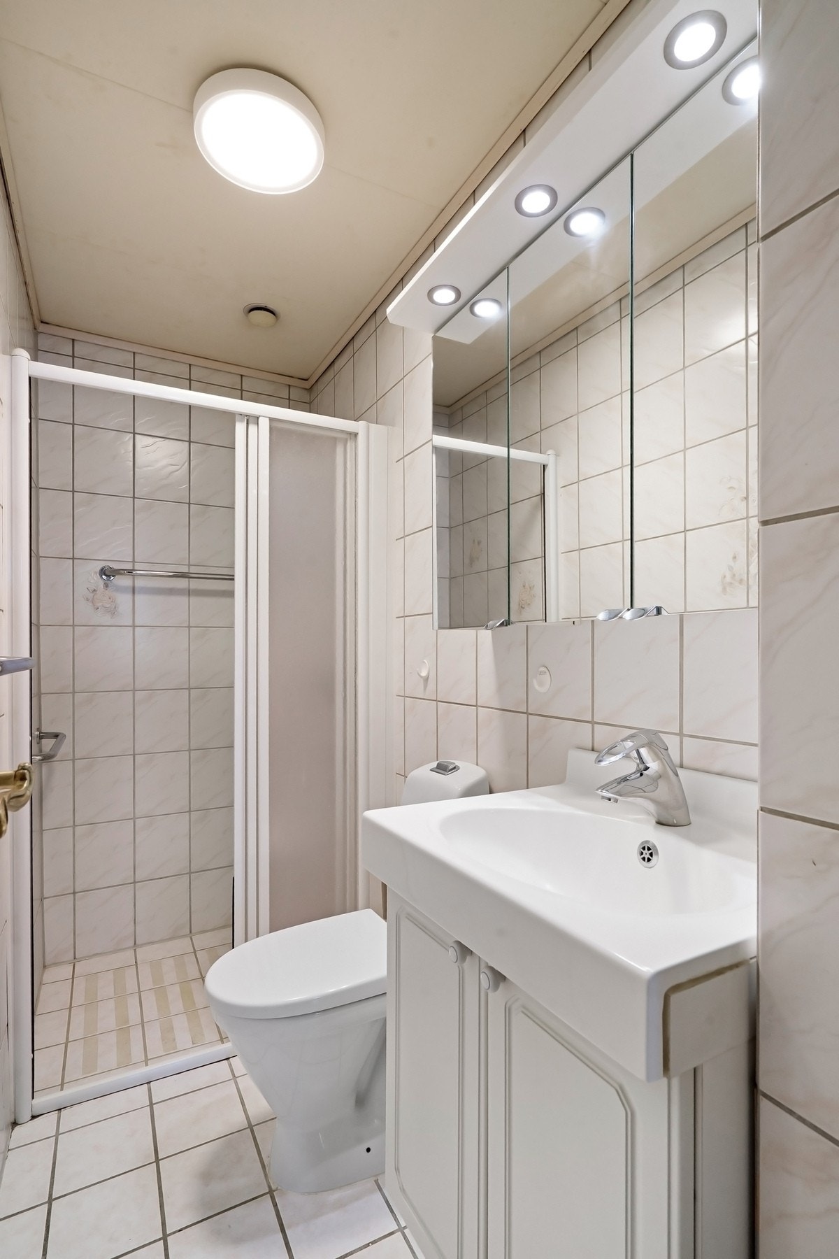 Flislagt baderom som består av benkeskap, veggskap med speilfronter over servant, gulvmontert toalett og dusjhjørne. Galleribilde